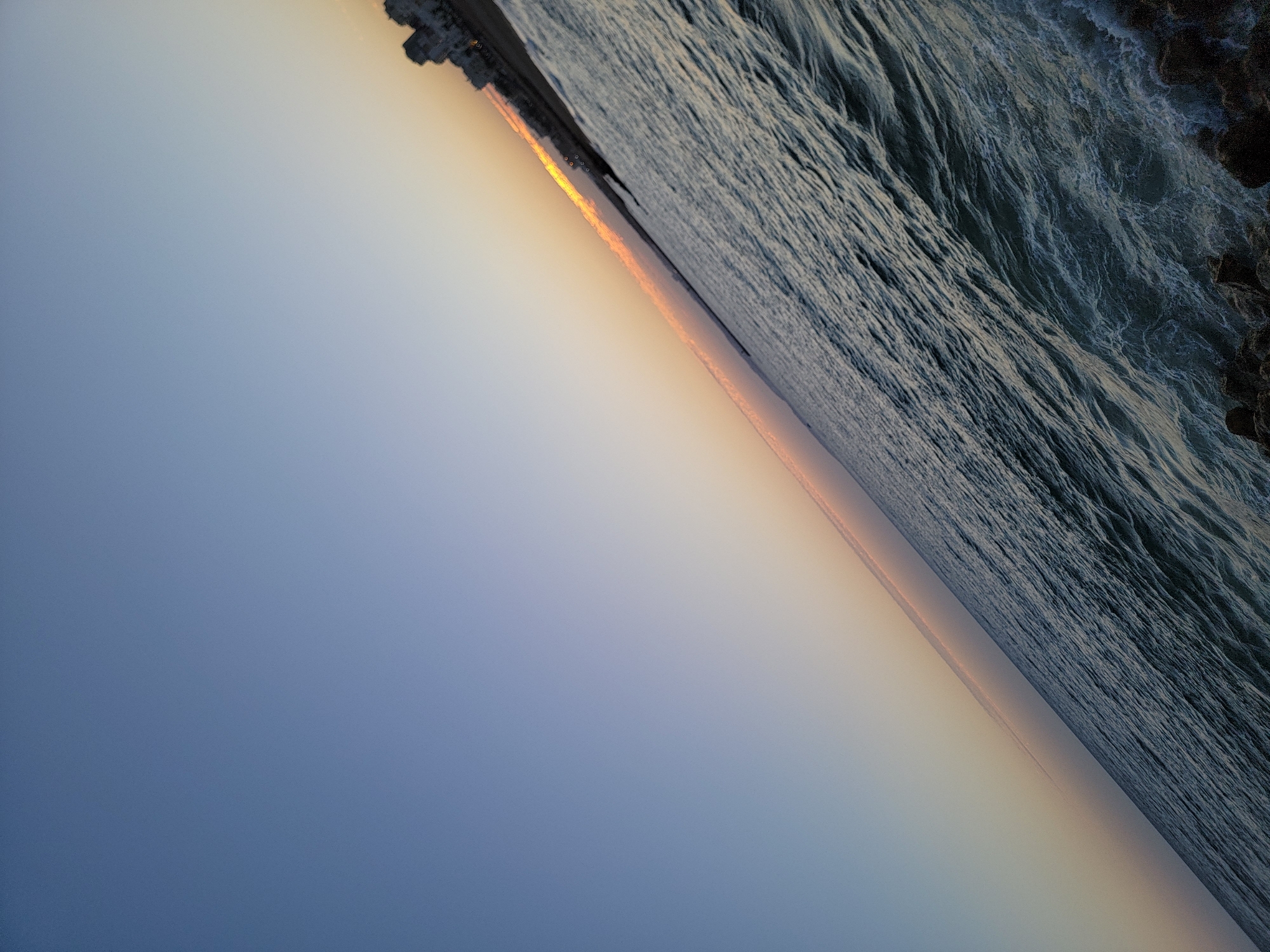 A diagonal, low-angle shot captures the turbulent waters of the Atlantic Ocean at sunset, viewed from the Quarteira coastline in Portugal. The sky transitions from a clear, pale blue at the top left to a soft orange and pink hue near the horizon, where a thin strip of clouds is visible. The sun's glare is reflected on the water's surface, highlighting the choppy waves and foamy whitecaps that crash against the rocky shore in the foreground. In the background, a row of buildings, silhouetted against the bright sky, lines the coast. The image is devoid of any people or animals.