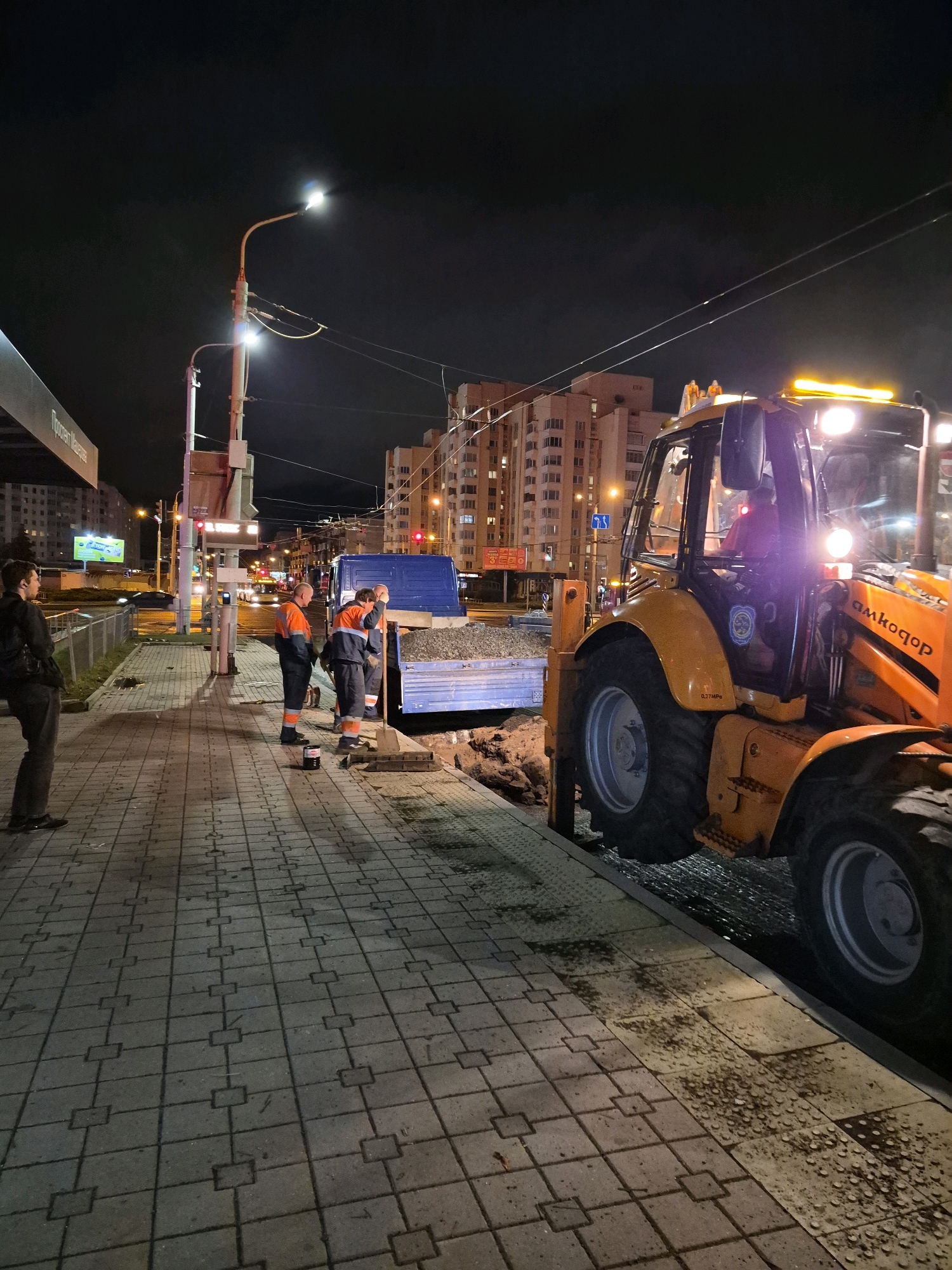 At night, on a sidewalk in Minsk, Belarus, construction workers in high-visibility vests are operating a yellow backhoe loader. The loader is positioned next to a trench dug into the sidewalk. A blue truck filled with gravel is parked nearby, and several workers are gathered around the trench, appearing to be engaged in some form of construction or repair work. The scene is illuminated by streetlights and the lights of the construction equipment. The background shows apartment buildings and streetlights, indicating an urban setting.