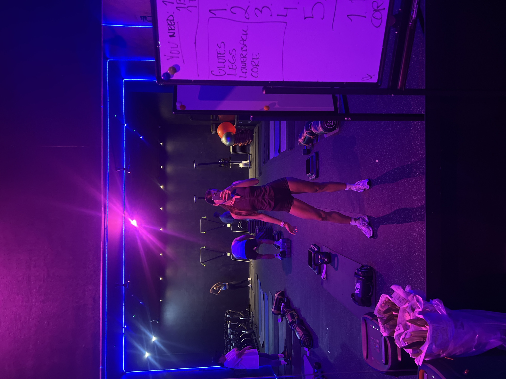 The image, oriented counter-clockwise, depicts an indoor fitness studio with dramatic purple and blue neon lighting, creating an energetic atmosphere. In the foreground, a woman wearing a dark top, shorts, white sneakers, and sunglasses on her head, is centered and appears to be posing or taking a selfie. She stands with her arms slightly bent and legs extended.
In the midground and background, several other individuals are engaged in various workout activities. One person is visible bent over an exercise machine, while others are further in the distance. The studio is equipped with various fitness gear, including weights, exercise machines, and medicine balls. A water bottle with "KS" written on it is visible near the floor.
A white board prominently displays workout instructions. Visible text on the board includes "YOU NEED: 1", a list of muscle groups: "GLUTES", "LEGS", "LOWER BACK", "CORE", and numbers such as "1", "1", and "12345". Another partial word, "COR", is also present. The dynamic lighting creates bright flares across the scene, illuminating the dark interior. In the bottom right corner, a clear plastic bag filled with what appears to be wads of money or papers is resting on a piece of equipment. The overall impression is of an intense group workout session at a modern gym.