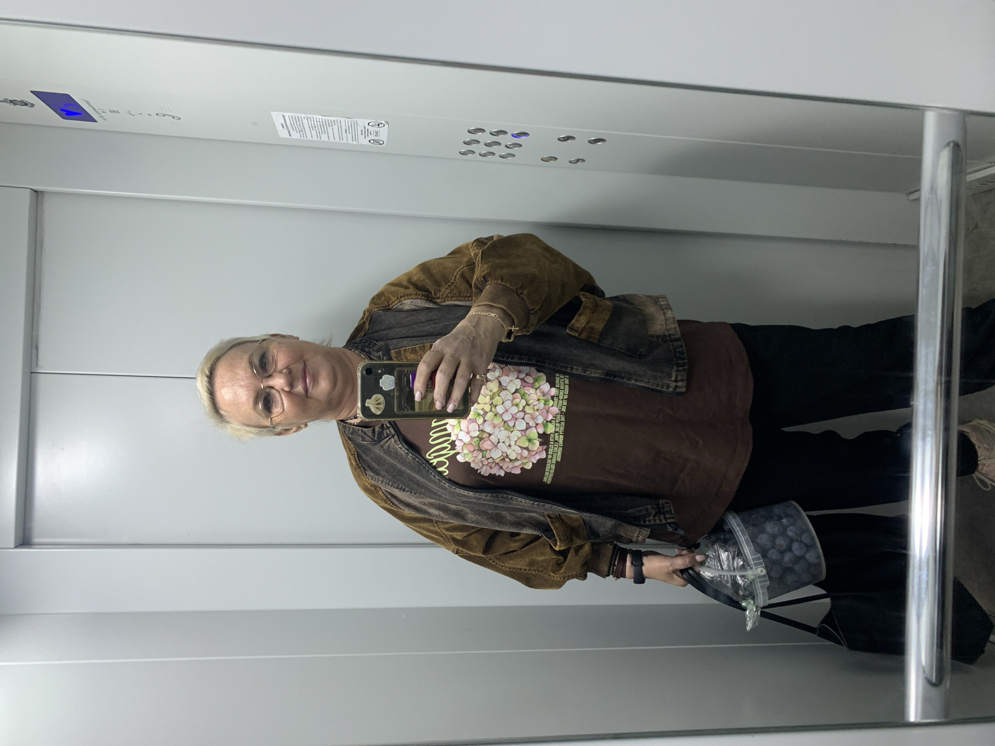 A woman with short blonde hair and glasses, wearing a brown jacket over a brown t-shirt with a floral graphic, stands in an elevator and takes a selfie. She is holding a clear plastic bucket filled with blueberries and a black tote bag in her left hand. The elevator walls are light gray, and a panel of buttons is visible on the right. The scene appears to be indoors, likely in a residential building in the city of Chaikovsky, Russia, given the context provided.