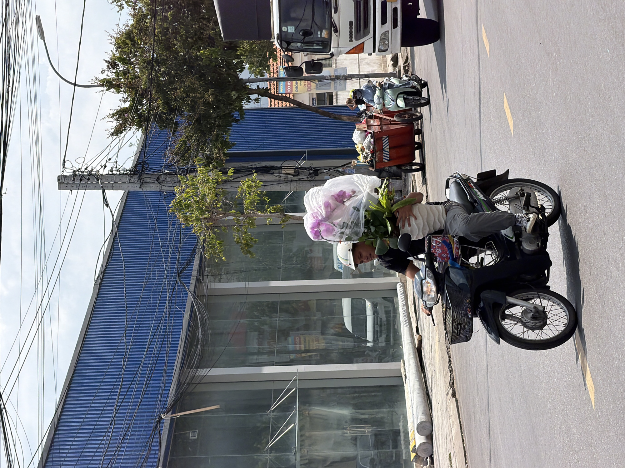 On a sunny day in Nha Trang, Vietnam, a man rides a black motorcycle down a paved road. He is wearing a white helmet and a black t-shirt, with gray pants and white sneakers. He is holding a large bouquet of pink flowers wrapped in plastic. In front of him, on the motorcycle, is a basket containing some items. Behind him, a woman on a light green scooter rides in the same direction. A red cart filled with trash is being pulled by the scooter. To the right, a white truck is parked on the side of the road. In the background, a blue building with a corrugated metal facade stands tall, with a large amount of electrical wires draped across it and the surrounding area. The street is marked with yellow lines and there are trees and streetlights visible.