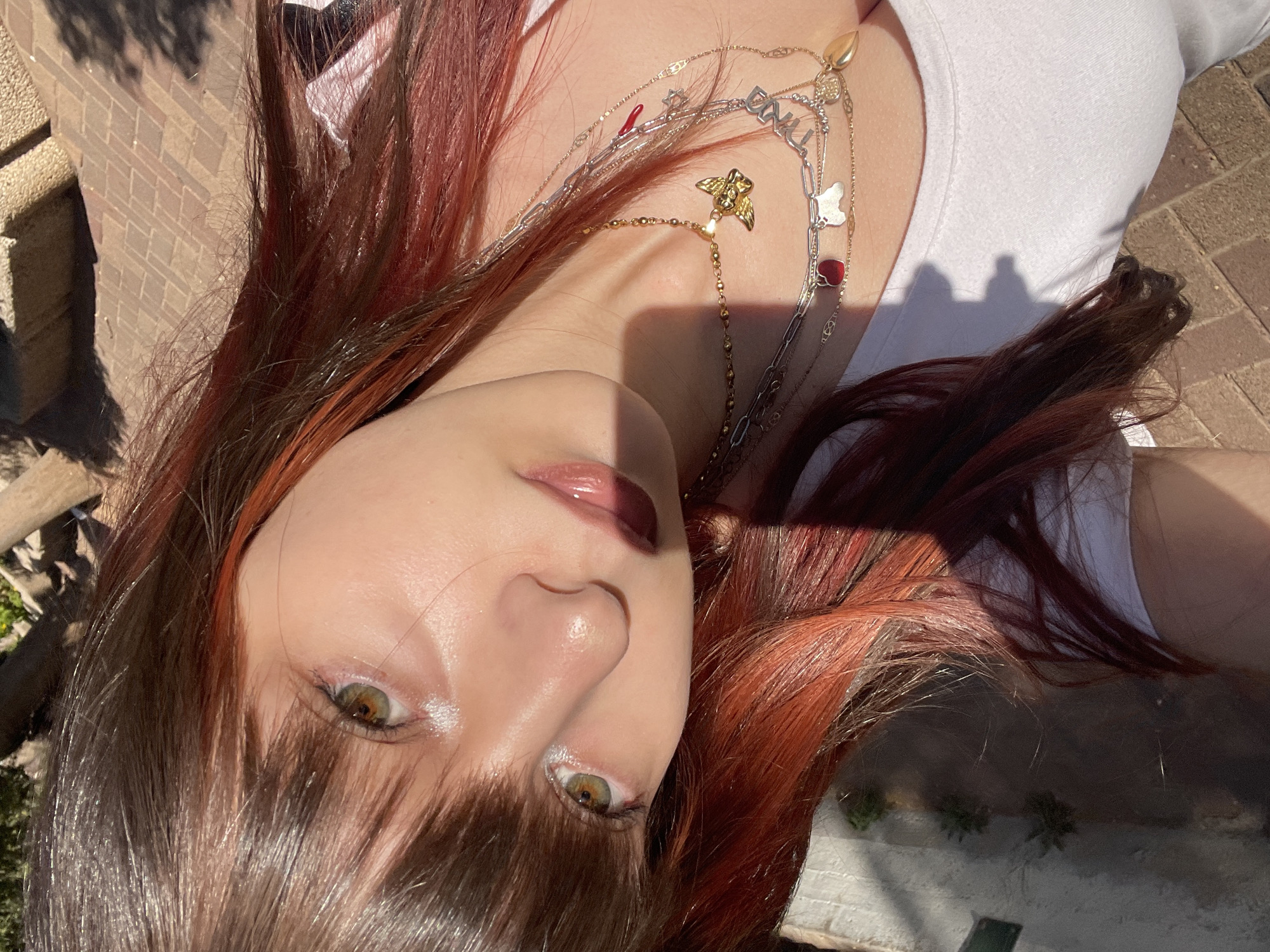 A young woman with brown hair with red highlights is pictured from a low angle, looking directly at the camera. She is wearing multiple necklaces, including a gold angel pendant, and a white top. The location appears to be outdoors in daylight, possibly in Raanana, Israel. The background shows a brick-paved area and some greenery, suggesting an urban or suburban setting. The overall composition is a close-up, emphasizing the subject's face and jewelry.