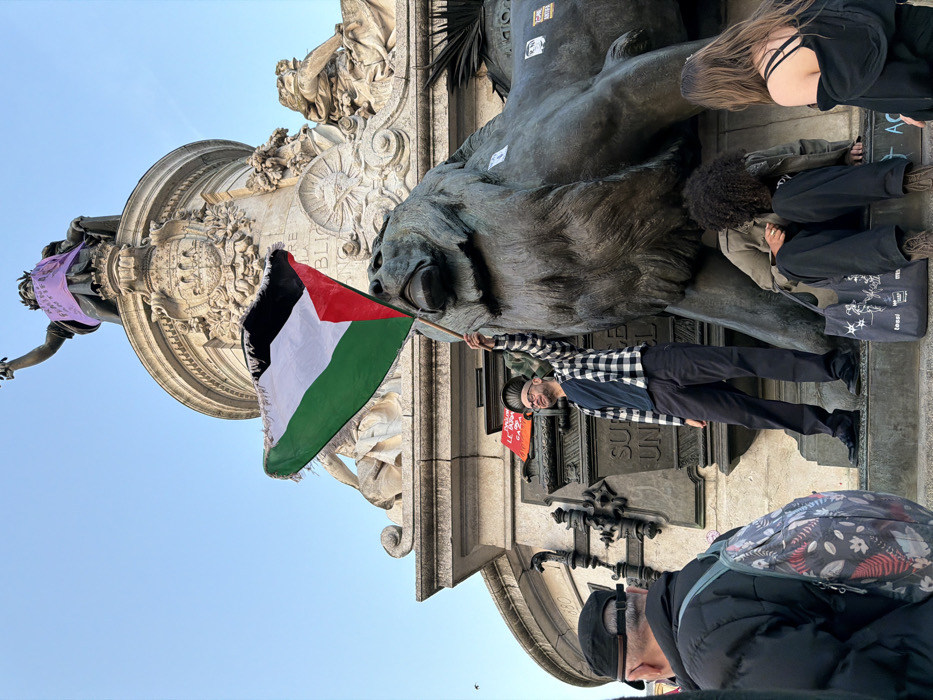 The image captures a scene at the base of the Monument à la République in Place de la République, Paris, France. The central feature is the monument itself, with a large bronze lion sculpture visible on the lower right. Above it, the monument's structure ascends, culminating in the statue of Marianne, which is adorned with a purple fabric banner displaying the partial text "FEMINISTES".
In the mid-ground, a male individual with short hair and glasses, wearing a black and white plaid shirt over a dark t-shirt and dark trousers, stands on a lower pedestal of the monument. He holds aloft a Palestinian flag, which is green, white, and black horizontally with a red triangle on the hoist side; its edges appear frayed. Behind this individual, a small red sign with partially visible French text, including "LE GAZA," is affixed to the monument.
To the right of the bronze lion, two individuals are partially visible: one person with dark, curly hair sits on the monument's base, clutching a dark tote bag with white designs; another person with long, dark hair stands beside them. In the immediate foreground, facing away from the camera, is a person wearing a black cap, a dark jacket, and a light-colored backpack adorned with a floral or leaf pattern. The scene is outdoors under a clear, light blue sky.
