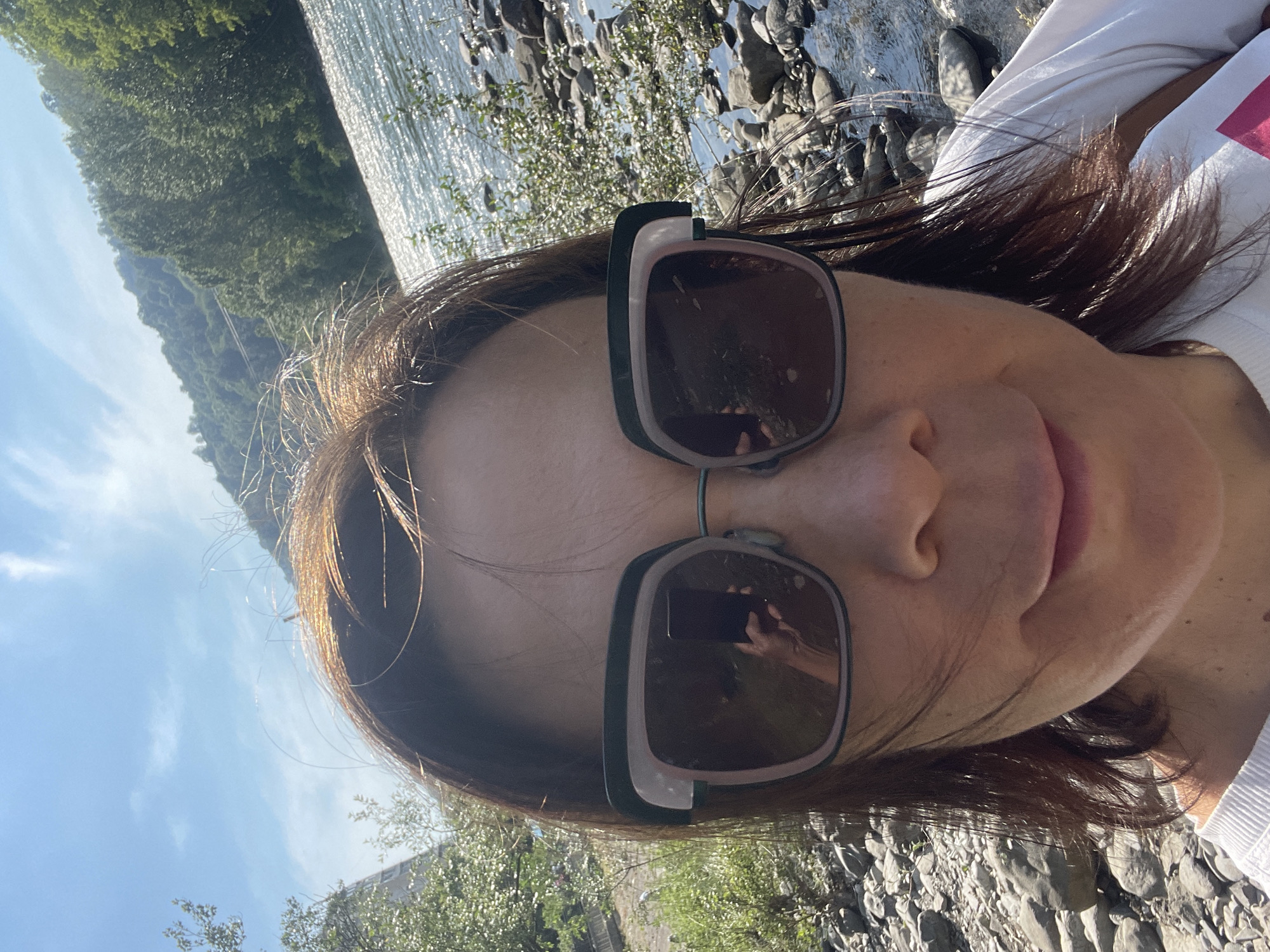The image is a close-up selfie of a woman outdoors, looking directly into the camera. She has shoulder-length, dark hair with visible lighter, possibly reddish-brown, strands on top, appearing somewhat disheveled. She wears large, dark-lensed sunglasses with a black and light pinkish-beige frame, obscuring most of her eyes. A reflection in her right sunglass lens (viewer's left) shows a hand holding a dark-colored smartphone, indicating she is taking the photograph. Her nose, mouth, and chin are visible, showing a slight, closed-lip smile. She is wearing a white top, part of which is visible on her shoulder and chest, with a hint of a pink design on the lower right side.
The background reveals a natural outdoor environment. To the viewer's right, a body of water, likely a river, flows with bright sunlight reflecting off its surface, creating shimmering patterns. The riverbank consists of numerous grey, rounded stones and rocks. Sparse green foliage and bushes grow along the water's edge. Beyond the river, a steep, densely forested hillside or mountain range rises, covered in lush green trees. To the viewer's left, more rocky ground similar to the riverbank is visible, with additional green vegetation and distant, partially obscured structures or buildings under a bright blue sky with scattered white clouds. The scene is illuminated by strong daylight.
Based on the combination of a wide river with rocky banks, and steep, heavily forested hillsides characteristic of the Caucasus Mountains, the location is consistent with the natural landscape found in and around Sochi, Russia, particularly along its major rivers.