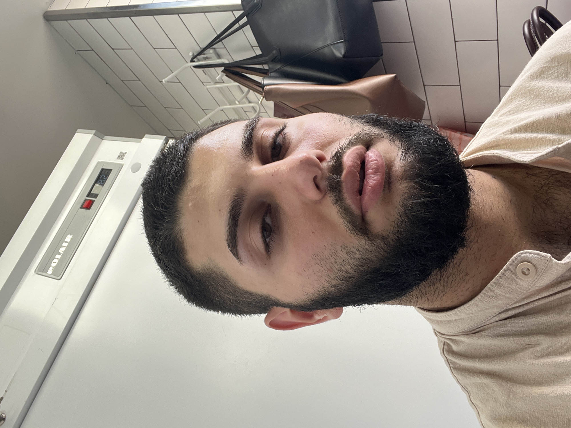 A close-up, waist-up, selfie-style portrait captures a young adult male with dark hair, a full beard, and dark eyes. His right eyebrow appears slightly swollen or discolored. He is wearing a light-colored, collared shirt with a button. The background features a white tiled wall with a silver rack holding two handbags, one black and one tan. To the left, a white refrigerator or freezer unit with a gray control panel displaying "POLAIR" and a digital readout of "82" is visible. The setting appears to be indoors, potentially in a kitchen or utility area. The image lacks any specific geographical markers to confirm its location within the city of Kommunarka, Russia.
