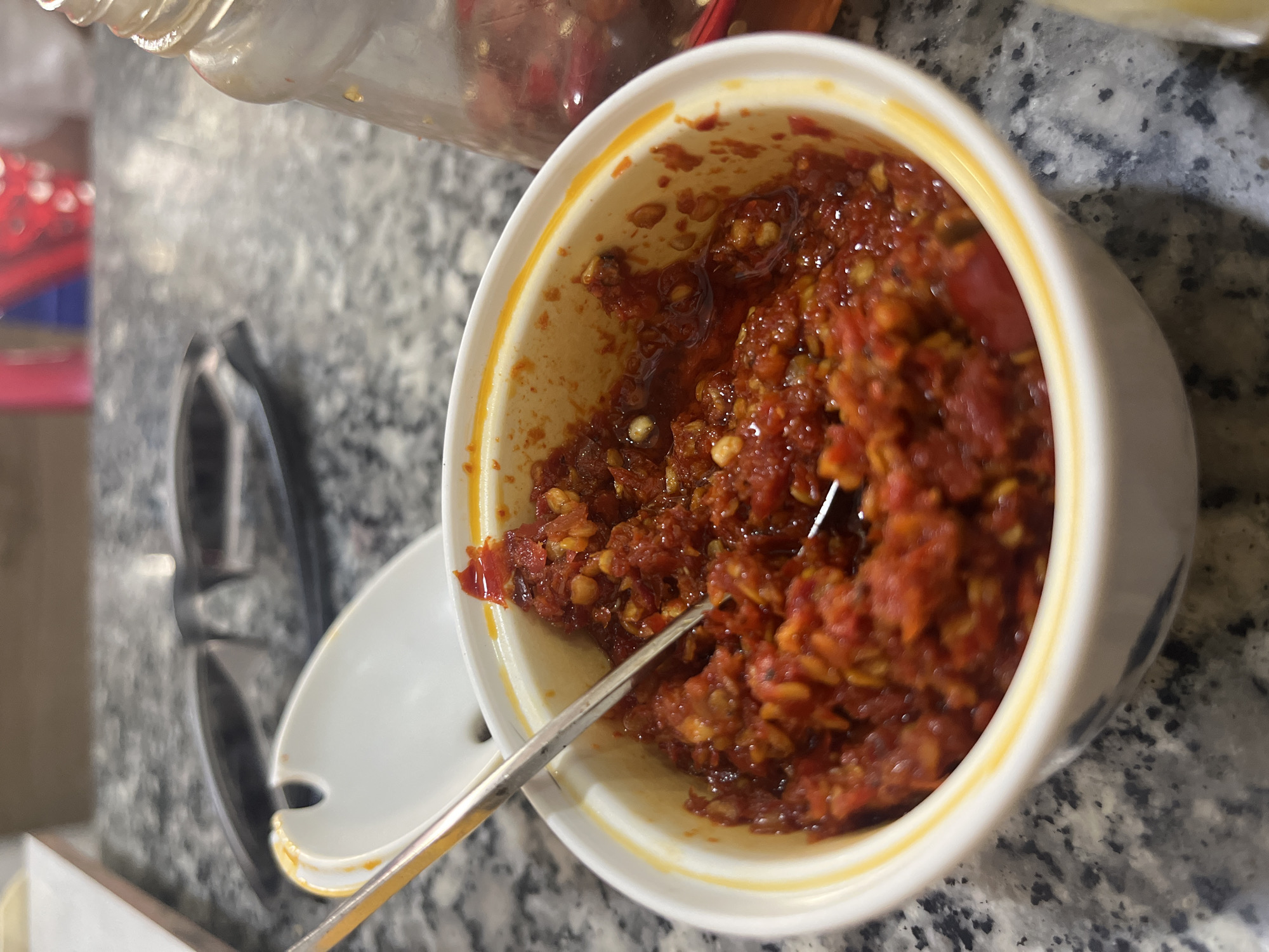 A close-up, slightly angled shot showcases a small, white ceramic bowl filled with a vibrant, red chili paste. The paste is chunky, appearing to be made from finely chopped chili peppers, possibly with oil and other seasonings mixed in. A silver spoon is partially submerged in the paste, suggesting it's a condiment. The bowl has a thin yellow rim and a subtle blue pattern around its base.
The bowl sits on a speckled gray countertop, likely granite. In the background, out of focus, a pair of black-rimmed glasses rests on the same surface. To the right of the bowl, a portion of a clear glass bottle is visible, containing red chili peppers in oil. The bottle has a textured cap. The overall impression is of a food-related setting, possibly a restaurant or a home dining area.
The specific location in the city of Bac Nha Trang Ward, Vietnam, cannot be determined from the visual information present in the image alone. This detail would require external context or metadata not provided.