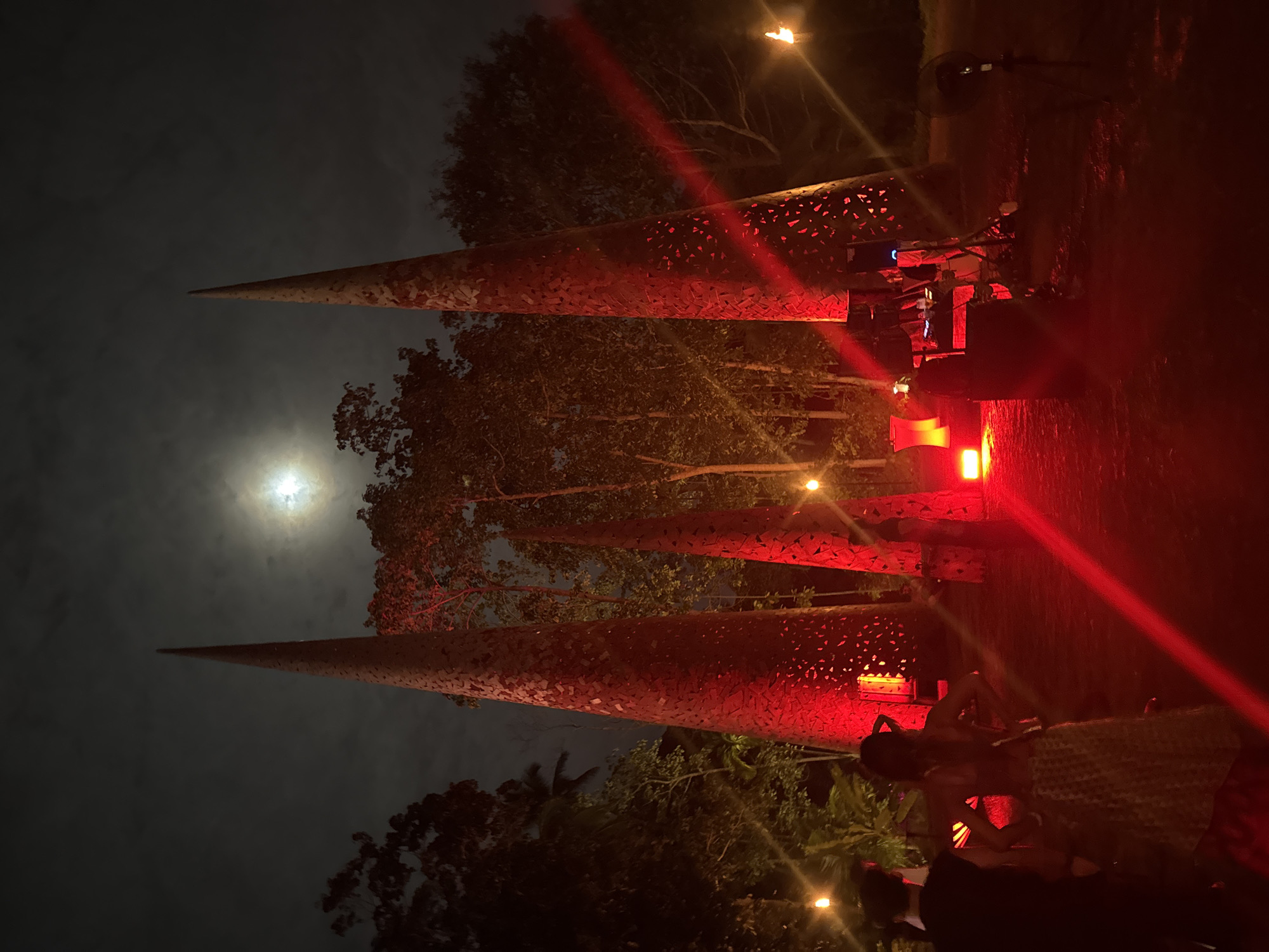 The image depicts an outdoor nighttime scene in Koh Samui District, Thailand, illuminated by a full moon and dramatic red lighting. Three tall, conical sculptures, each with a textured surface and cut-out patterns, dominate the foreground. The sculptures are lit from below with intense red light, casting a vibrant glow.
In the left foreground, two individuals are visible from the back. The person on the left is wearing dark clothing, and the person on the right is wearing a patterned skirt and a top. They appear to be observing the illuminated sculptures.
The environment is a tropical setting with dense foliage and trees surrounding the sculptures. The sky is cloudy, with the bright disc of the moon peeking through. To the right of the sculptures, a DJ booth or sound system setup is present, indicated by speakers and equipment. Further to the right, a traditional torch with a flickering flame provides additional illumination. Other red-lit objects, possibly seating or decorative elements, are visible in the mid-ground. The overall atmosphere suggests an event or gathering taking place amidst an artistic installation.