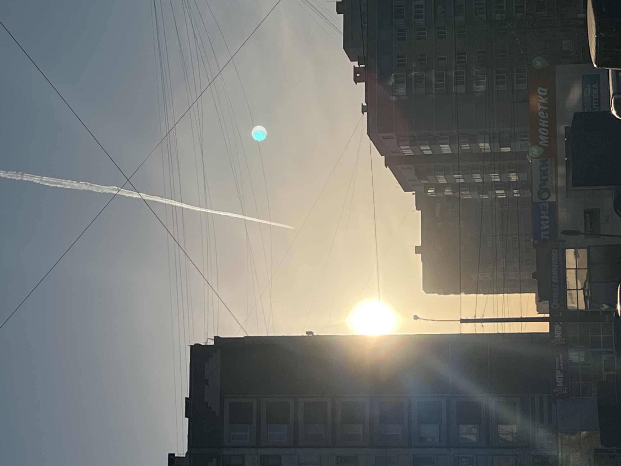 The image shows a cityscape in Izhevsk, Russia, at dawn or dusk. Several tall, multi-story buildings frame the scene. A bright, diffused sun is visible between the buildings, creating a strong backlight. Utility lines crisscross the sky. A contrail cuts across the upper left portion of the sky. A cyan orb-like object is also visible in the upper portion of the sky. Signage in Russian is visible on one of the buildings.