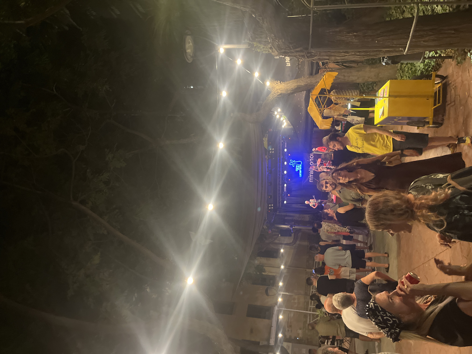 The image depicts an outdoor event at night, possibly a festival or concert, taking place in the city of Tarragona, Spain, specifically at "La Palmira," as indicated by signage. A band is performing on a stage illuminated by blue and white lights. A crowd of people stands before the stage, some watching the performance, while others are engaged in conversation. In the foreground, two women are conversing, one of whom is holding a red can. To the right of the stage, a yellow vending cart with an umbrella is visible. The scene is set amidst trees and buildings, with strings of lights overhead creating a festive atmosphere.