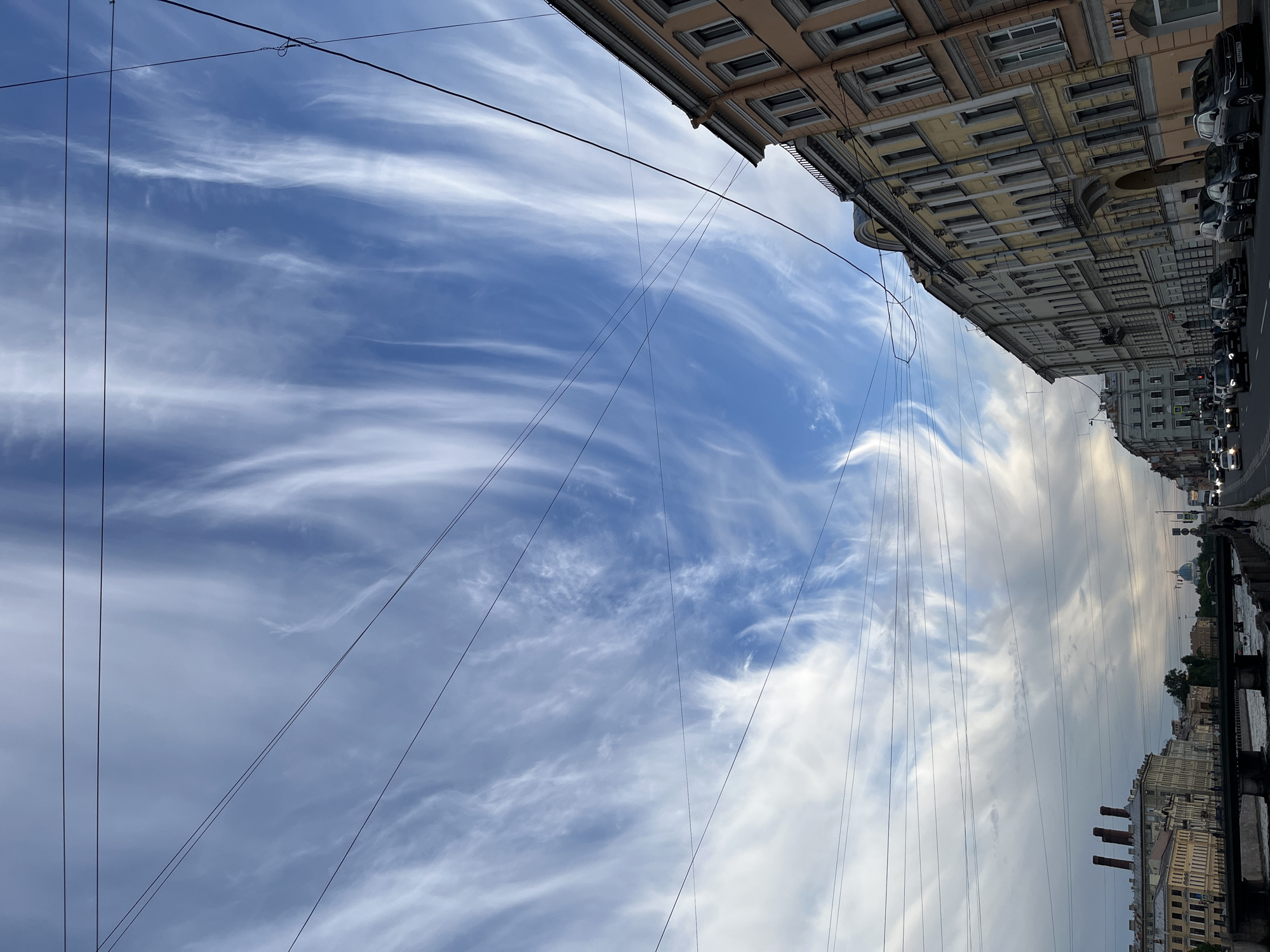 The image, oriented correctly, presents an upward-looking view of a city sky dominated by streaky white clouds against a bright blue background. Numerous thin black utility wires crisscross the sky, some running horizontally and others diagonally across the frame.
On the right side of the image, a row of multi-story classical buildings lines a street. These buildings feature multiple windows, cornices, and decorative elements, appearing in shades of pale yellow, peach, and gray. Several dark-colored cars are parked or moving along the street at the base of these buildings.
In the bottom right corner, a body of water, likely a canal, is visible, reflecting the sky. Across the canal, a distant cityscape can be seen, including a prominent blue dome, possibly belonging to a church or cathedral, characteristic of Saint Petersburg. Three dark, cylindrical smokestacks are also visible on a building near the canal.
The time of day appears to be daytime, possibly late morning or afternoon, indicated by the bright, clear sky on the left transitioning to slightly more overcast conditions on the right. The weather is partly cloudy with wispy, high-altitude clouds. The architectural style, presence of canals, and the distant blue dome are strong visual cues aligning with Saint Petersburg, Russia. No visible text is discernible in the image.