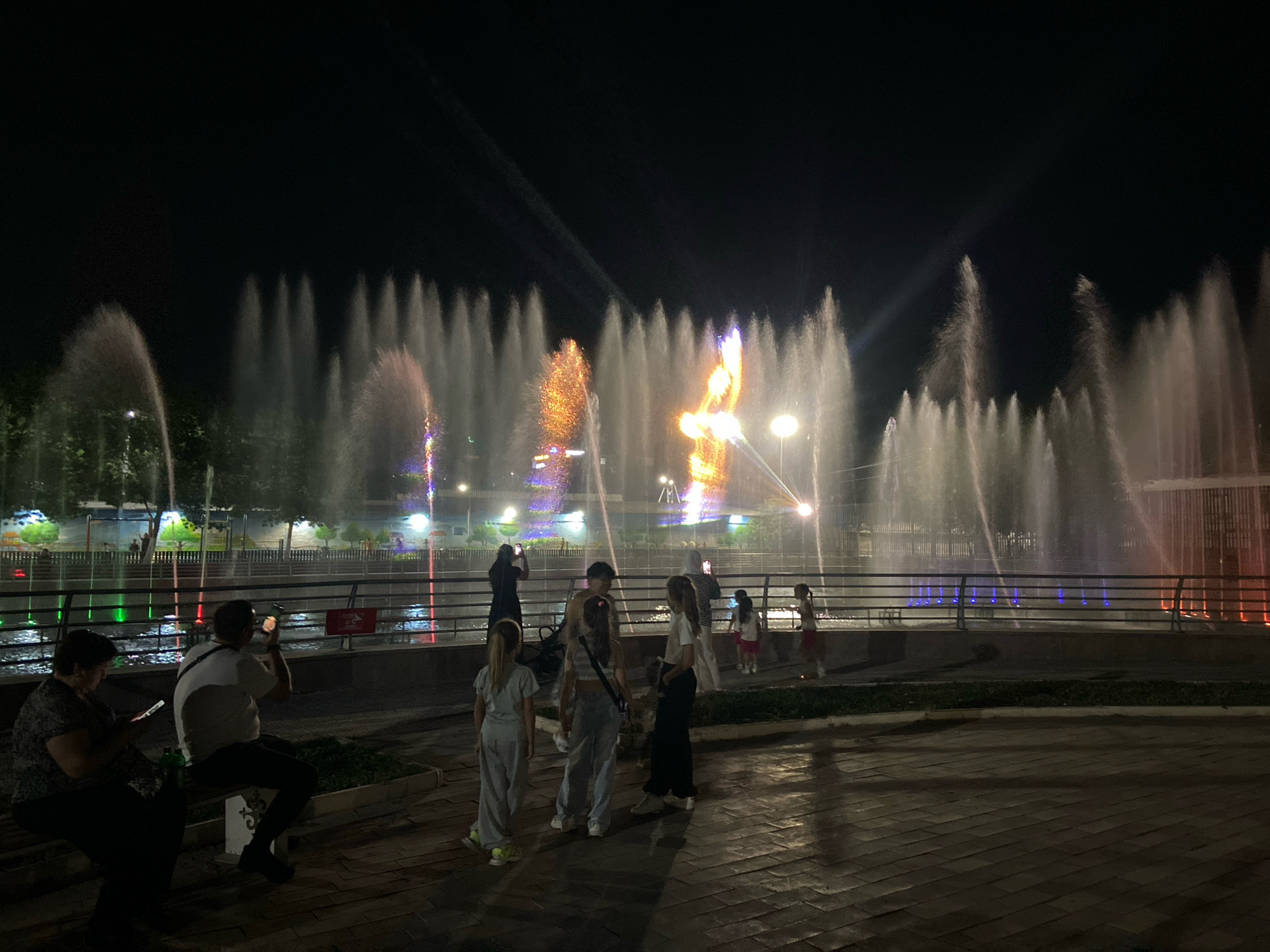 The image shows a nighttime view of a large, illuminated musical fountain in Shymkent, Kazakhstan. The fountain is the central focus, with numerous water jets spraying upwards, creating a dynamic visual display. Some jets are illuminated with colorful lights, including orange, purple, and green, adding to the spectacle.
In the foreground, several people are visible. On the left, an older woman sits on a bench, looking at her phone. Next to her, a man is seated and appears to be taking a photo with his phone. Further to the right, a group of younger individuals, likely children and teenagers, are standing and observing the fountain. One girl in a light grey outfit is facing away from the camera, while another in a striped top and blue jeans is turned slightly to the side. Another young person in dark clothing is also present.
The background features trees and buildings behind the fountain, partially obscured by the water and lights. The ground in the foreground is paved with bricks, and a grassy area is visible between the paved area and the fountain's edge. A railing separates the viewing area from the fountain. The overall atmosphere is one of evening entertainment and public gathering around a prominent city attraction.