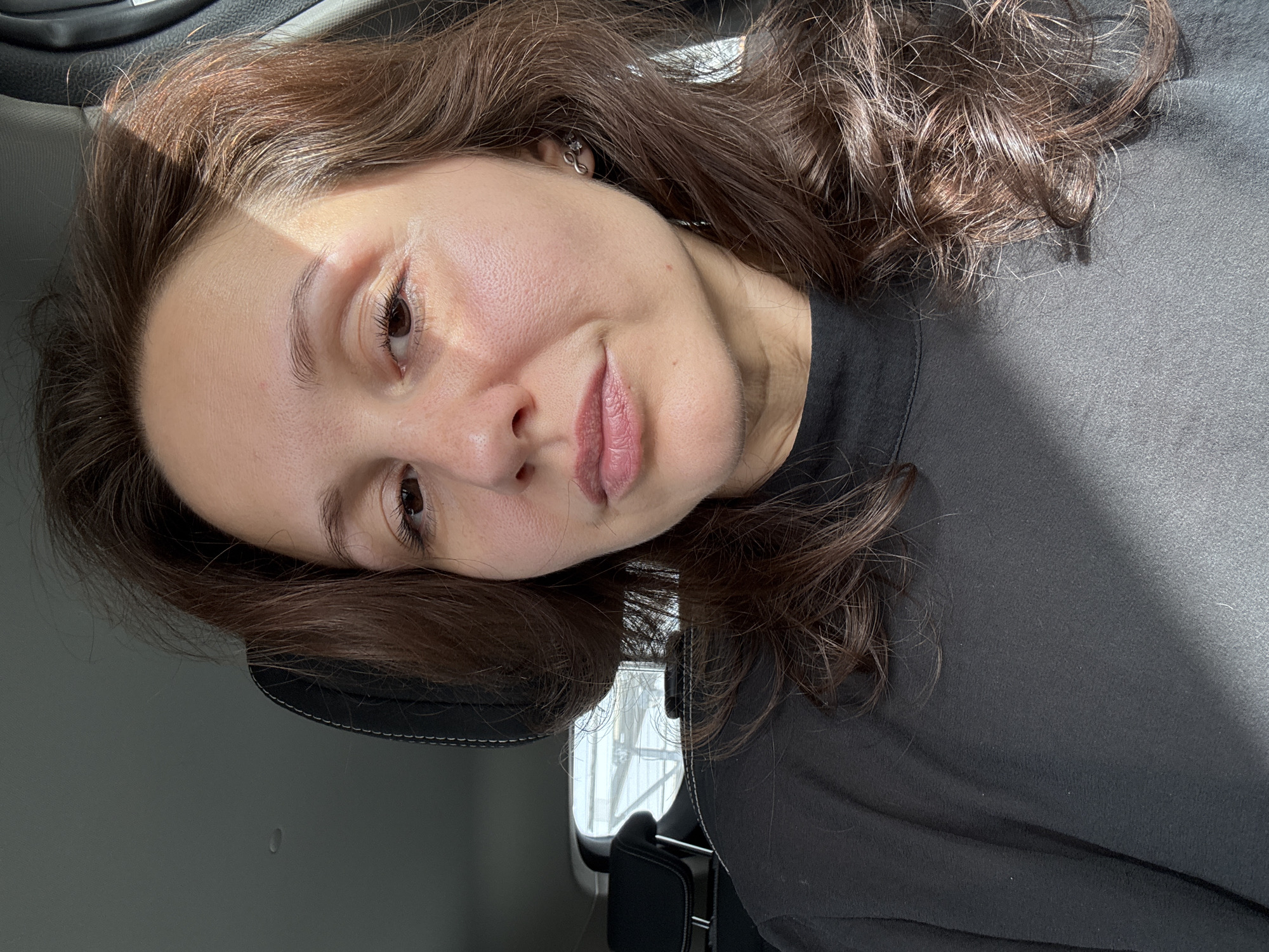 A woman with long, wavy brown hair is seated inside a vehicle. She is wearing a dark gray or black top. The woman is resting her head on the headrest. Sunlight illuminates part of her face. The image's background shows a blurred view outside the vehicle window. No information suggests the location is Dubai, United Arab Emirates.
