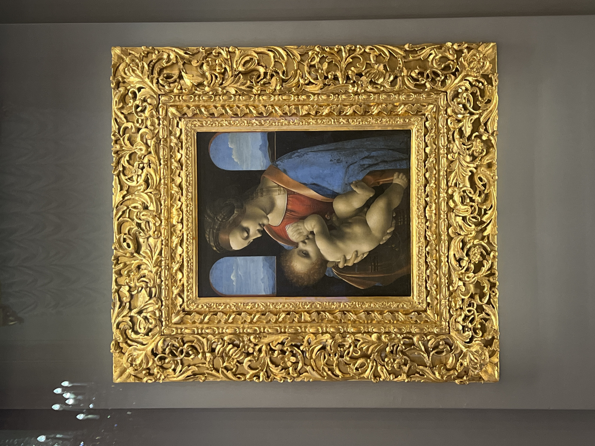 The image shows a painting of the Virgin Mary and baby Jesus in an ornate gold frame. The painting is oriented sideways. The background of the painting is dark with two arched windows showing a blue sky. Mary is wearing a blue robe and a red dress, and she is holding the baby Jesus. The frame is highly detailed with a floral design. The painting is hanging on a gray wall. The location appears to be in a museum or art gallery in the city of Unknown, Unknown.