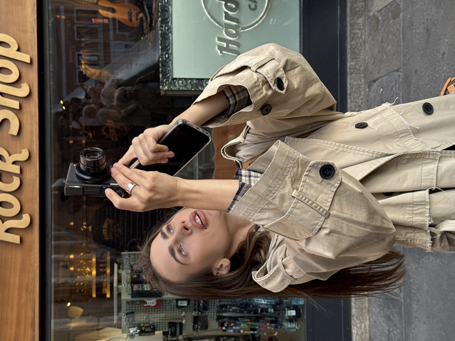 A female subject is depicted on an urban street in Venice, Italy, positioned in front of a Hard Rock Cafe and Rock Shop. She has long brown hair and wears a light-colored trench coat over a plaid shirt, with a silver ring on her right ring finger. With both hands raised, she holds a black compact digital camera with her right hand and a black smartphone in a clear case with her left hand, both devices pointed upwards. Her head is tilted back, and her gaze follows the upward trajectory of the devices. Behind her, a large glass shop window features "Rock Shop" in gold lettering above and "Hard Rock Cafe" in silver lettering on the lower right. The window reflects street activity and displays merchandise, including guitars. The ground is paved with stone.