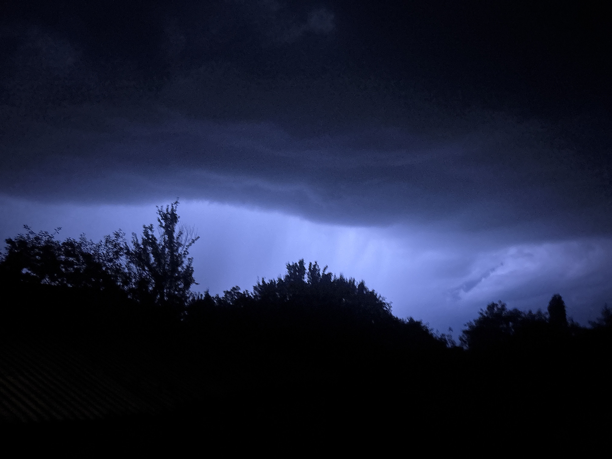 The image depicts a nocturnal or severely overcast outdoor scene dominated by dark, dense storm clouds. A broad horizontal band of intense electric blue and purple light illuminates the lower cloud layers across the mid-ground, suggesting an internal lightning flash or strong illumination source behind the clouds. The upper sky is deep indigo to black, with subtle texture in the cloud formations. In the foreground, a continuous, undulating silhouette of trees and dense foliage spans the lower portion of the frame, appearing entirely black against the brighter sky. A faint, dark, horizontally ribbed surface, possibly a roofline or fence, is visible in the extreme lower-left corner, also in deep shadow. No human figures, animals, or specific identifiable man-made structures are visible. The specific location in the city of Unknown, Unknown is not discernible from the image.