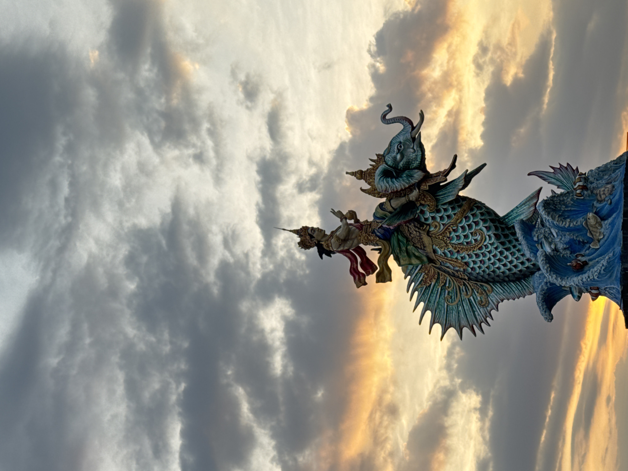 The image captures a low-angle shot of a Hindu temple architectural element in Mengwi, Indonesia, set against a dramatic sky. The sky displays a gradient of colors, ranging from dark gray clouds on the left to golden-yellow hues on the right, suggestive of either sunrise or sunset.
The temple element features a complex sculpture with an elephant-headed figure, intricate ornamentation, and scale-like textures, possibly representing a mythical creature. Smaller human figures are integrated into the design. The structure is painted in vibrant blues, greens, and golds, contrasting with the sky's muted tones.