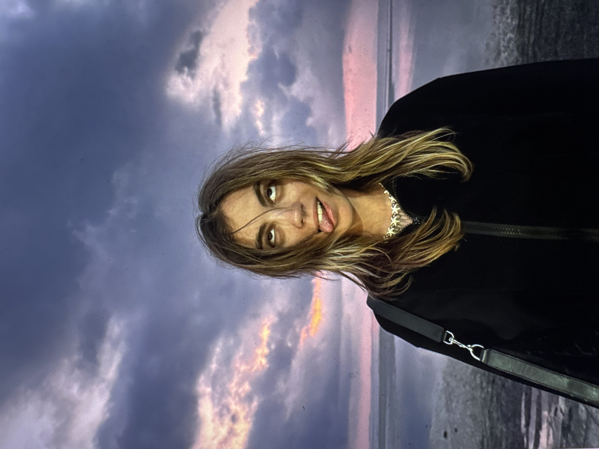 A young person with long, blonde-streaked brown hair stands outside during twilight. They are wearing a black zippered hoodie and a necklace with a floral-like design. The person is sticking their tongue out and rolling their eyes upwards, creating a playful or humorous expression. A dark bag is slung over their left shoulder. The background features a dramatic sky filled with purple and pink clouds, indicative of a sunset or sunrise, over a body of water and a dark, sandy shore. The scene appears to be a natural, outdoor setting, likely a beach or waterfront. There is no indication that this location is in Moscow, Russia.
