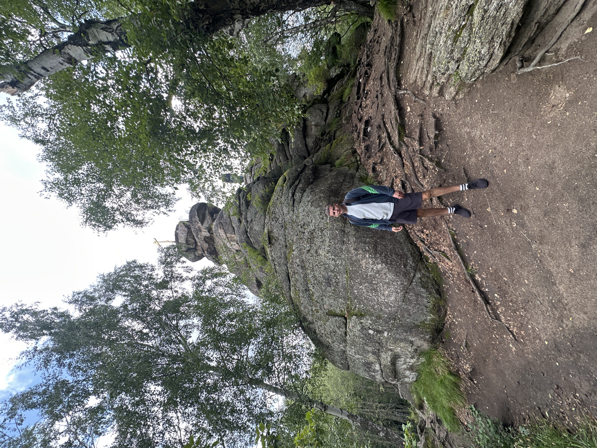 A man stands on a dirt path in a forest at the foot of a large rock formation. The man is wearing a gray t-shirt, a dark blue and green jacket, dark shorts, black athletic shoes, and white-striped socks. He has a backpack on his back. The rock formation is made up of large, grey boulders, some covered in moss. A golden cross is visible at the top of the rock formation, suggesting a religious significance to the location. Trees with green leaves surround the area, and the sky is cloudy. The path is covered with dirt, leaves, and exposed tree roots. The image appears to have been taken from a low angle, looking up at the man and the rock formation. The overall setting suggests a natural, outdoor environment, likely a hiking trail or a tourist attraction. The location is identified as being in the city of Belokurikha, Russia.