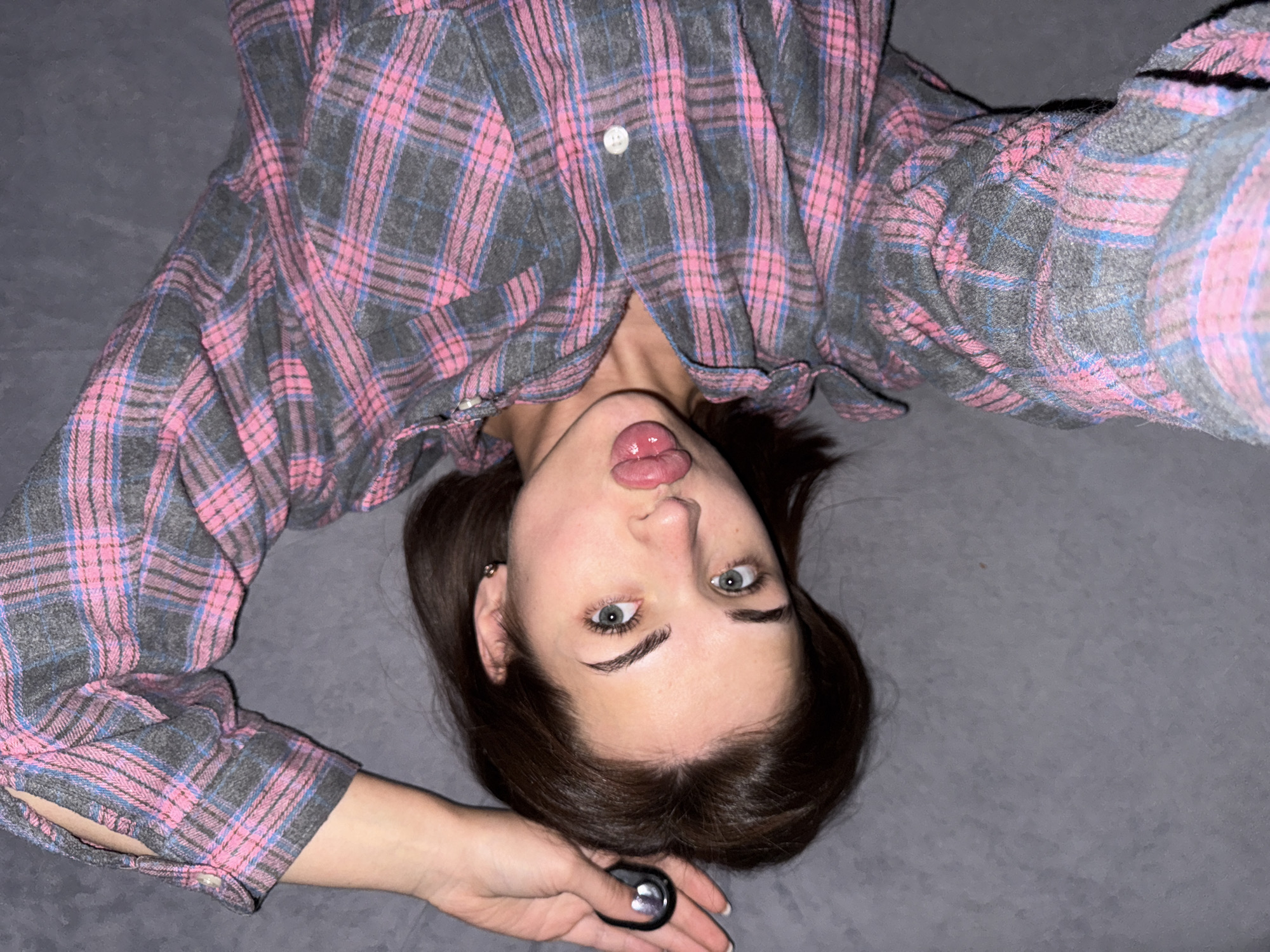 A woman lies on a gray surface, possibly a bed or couch, wearing a pink and gray plaid shirt. She's upside down relative to the viewer and sticking her tongue out. She is holding a small, dark object in her hand. The location is not discernible from the image.