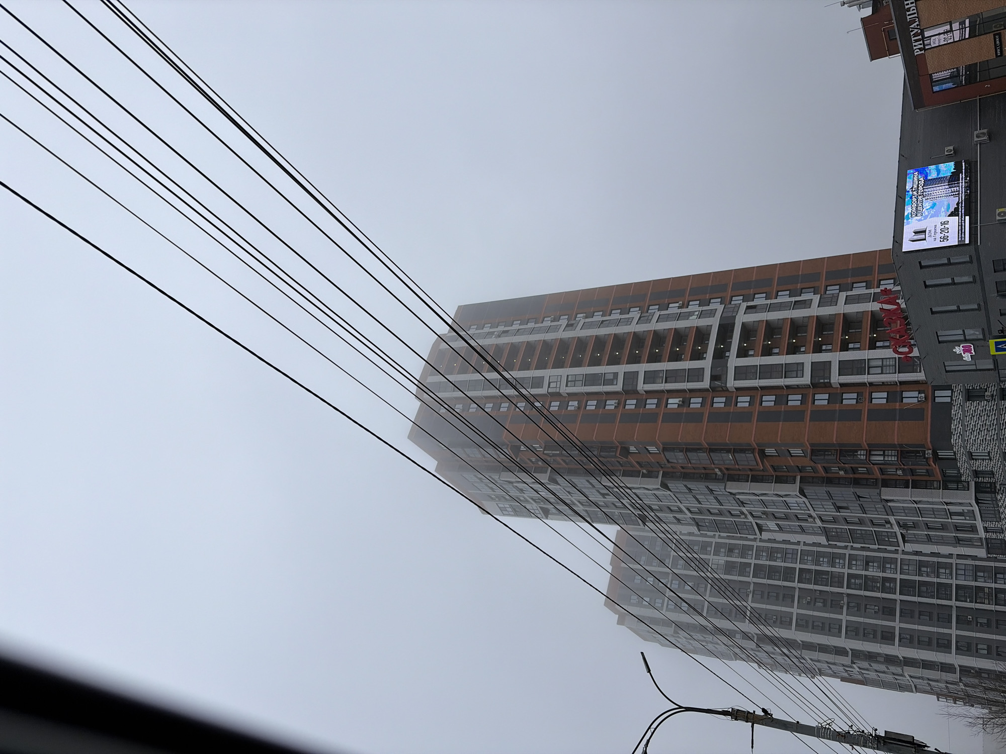 The image shows a street scene in Izhevsk, Russia, characterized by a heavy fog obscuring the view. A modern high-rise building dominates the frame, its façade partially visible through the mist. The building has alternating sections of grey and orange/brown coloring. A sign on an adjacent building advertises "ΔΟΜ на Герцена" (house on Herzen), with the text "КОМФОРТ, И ТИШИНА В ЦЕНТРЕ ГОРОДА" (comfort and silence in the city center) and a phone number: 56-20-16. Other nearby signs read "РИТУАЛЬН" and "КРИСТ, АДВОКАТ". Power lines crisscross the sky.