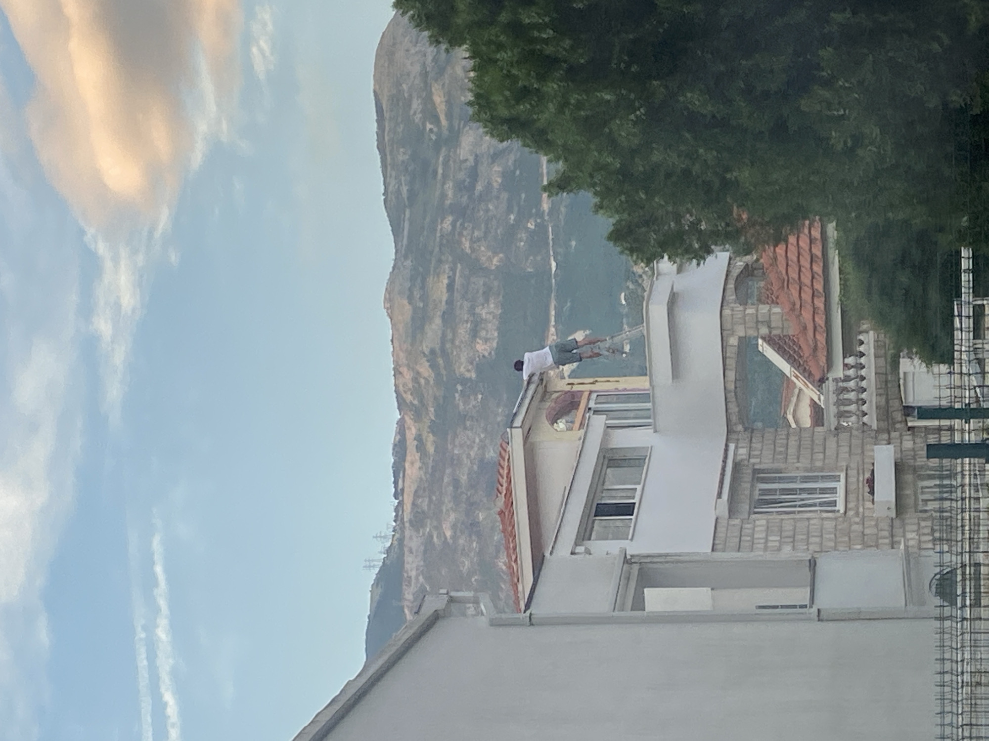 A medium shot depicts a person standing on a rooftop of a building with a ladder, against a backdrop of a mountain and a partly cloudy sky. The building is located in a residential area of Budva, Montenegro. The building's architecture includes features like red tile roofs, windows, and brickwork. The person on the roof is wearing light-colored clothing. The surrounding environment includes trees and other buildings, suggesting a densely populated area. There is an electrical grid on the side of the mountain.