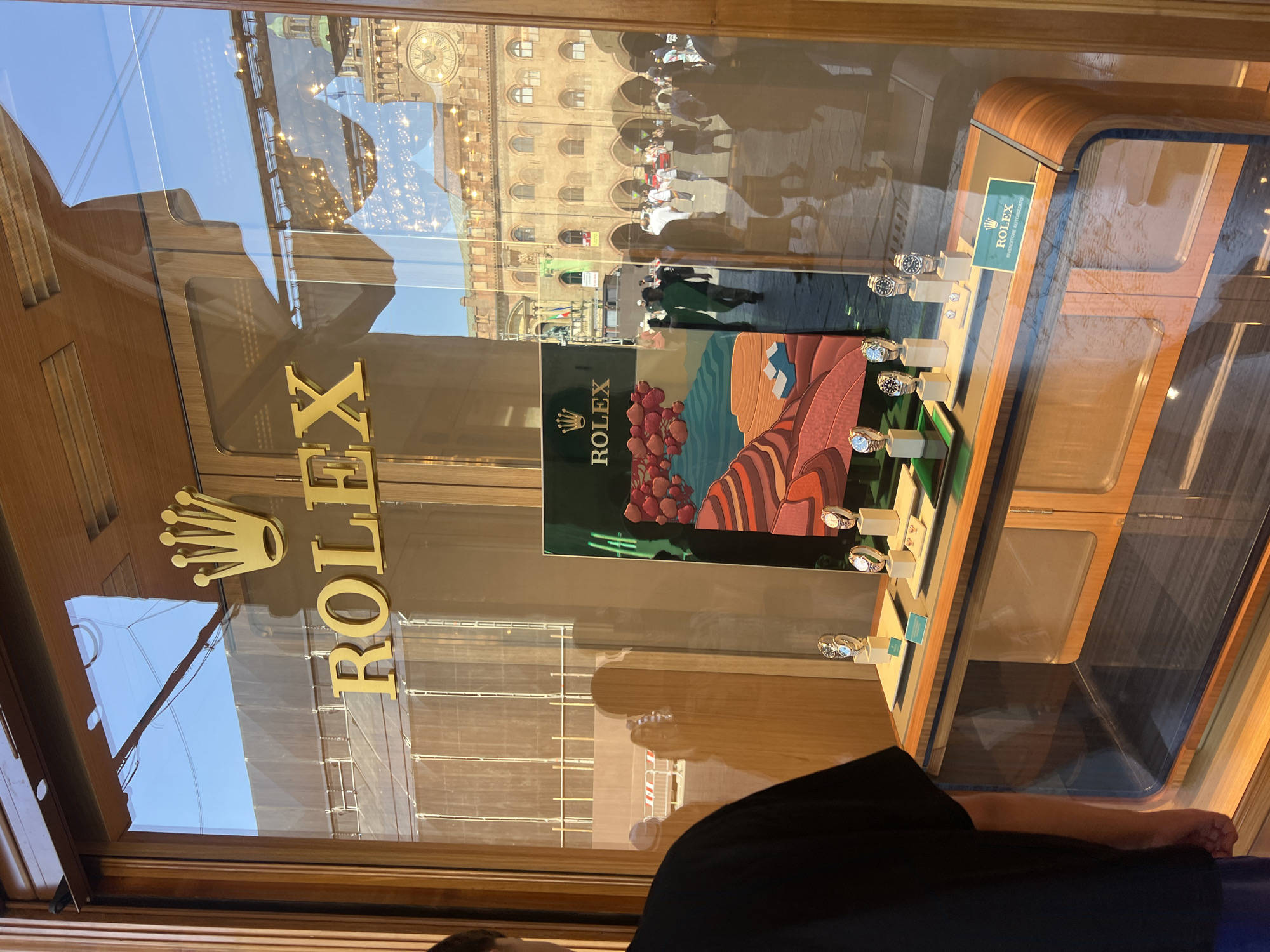 The image depicts a Rolex store window in an urban setting. The gold Rolex logo is prominently displayed on the glass. Inside the window, several Rolex watches are showcased on pedestals. A Rolex advertisement with a landscape design is also visible. The reflection in the window reveals a building with a clock tower and several people walking by. The building has arches. The overall scene suggests a high-end retail location in a city with historical architecture.