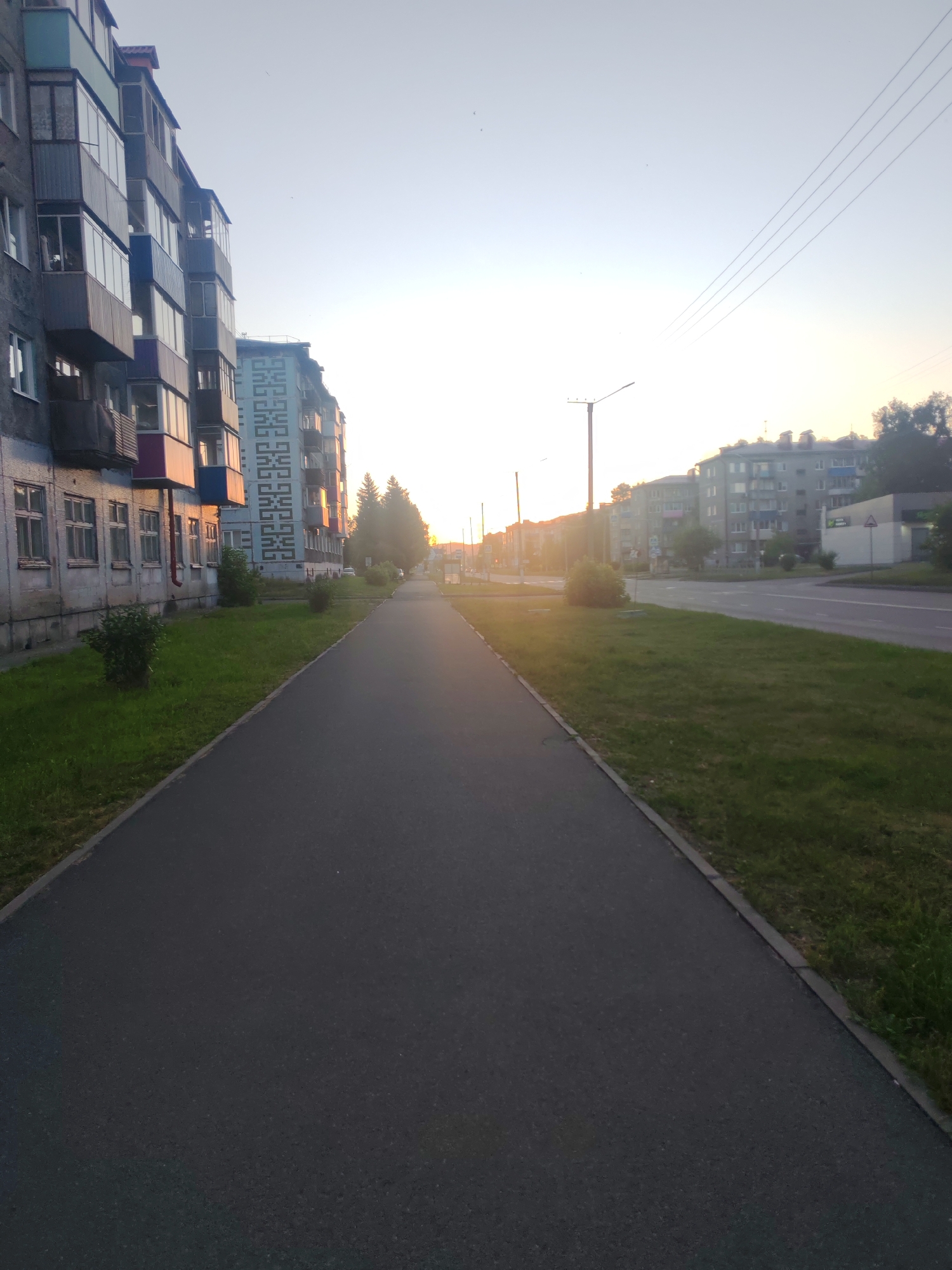A street scene in Mezhdurechensk, Russia, at what appears to be dusk or dawn. A paved pedestrian walkway runs diagonally from the bottom left to the top right of the frame, bordered by a strip of green grass on the left. Adjacent to the grass is a weathered, multi-story residential building with multiple balconies, some of which are enclosed. The building's facade shows signs of age. Further down the street, on the right side of the walkway, another residential building with a lighter-colored facade featuring a repeating geometric pattern is visible. Beyond that, more multi-story apartment buildings form the cityscape, with trees and a road also present. Overhead, power lines are strung across the pale sky, and a street lamp stands to the left of the walkway. The light suggests either sunrise or sunset, casting a soft glow on the scene. There are no people or vehicles immediately visible in the frame.
