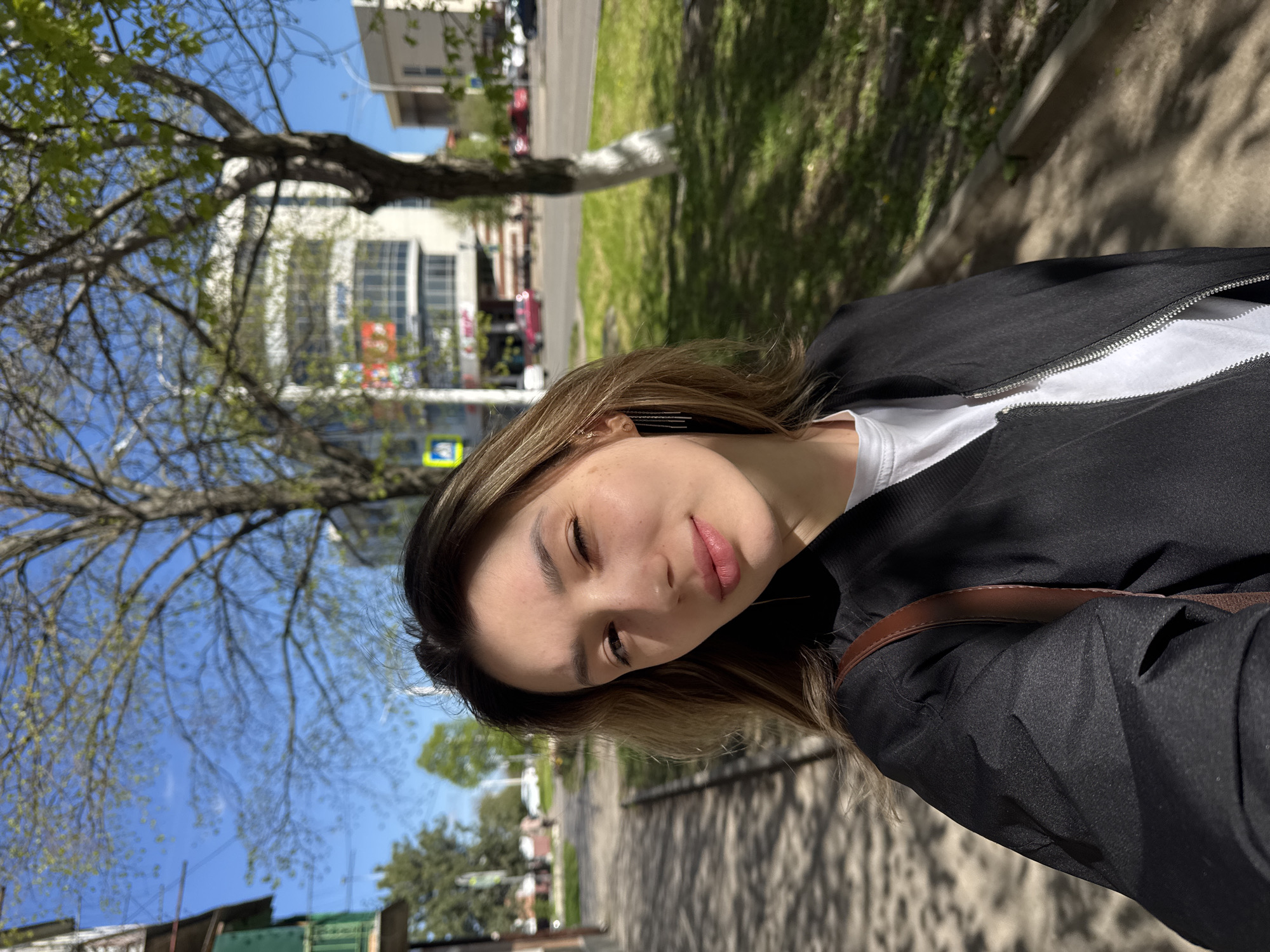 This image features a young woman taking a selfie outdoors on a sunny day. The photograph is rotated 90 degrees counter-clockwise.
The primary subject is a young woman with fair skin, light freckles, and medium-length brown hair with lighter highlights. She has a relaxed expression, with her lips slightly parted and her right eye partially closed, possibly winking or squinting in the sun. She is wearing a black zip-up jacket over a white t-shirt, and a brown strap, likely from a shoulder bag, is visible over her right shoulder.
The setting appears to be a paved path or sidewalk next to a grassy area, possibly a park or street in an urban environment. The background shows a clear blue sky and abundant natural light, indicating daytime. Trees line the path, some with bare branches and others beginning to show green leaves, suggesting spring. Distinct shadows are cast on the ground by the trees, further emphasizing the bright sunlight. In the mid-ground, several buildings of varying sizes are visible, along with glimpses of traffic or parked cars. A blue and yellow sign, possibly a road or pedestrian crossing sign, is partially visible behind her head.
The activity taking place is a casual portrait or selfie. There is no clearly legible text visible on any signs or buildings in the background.