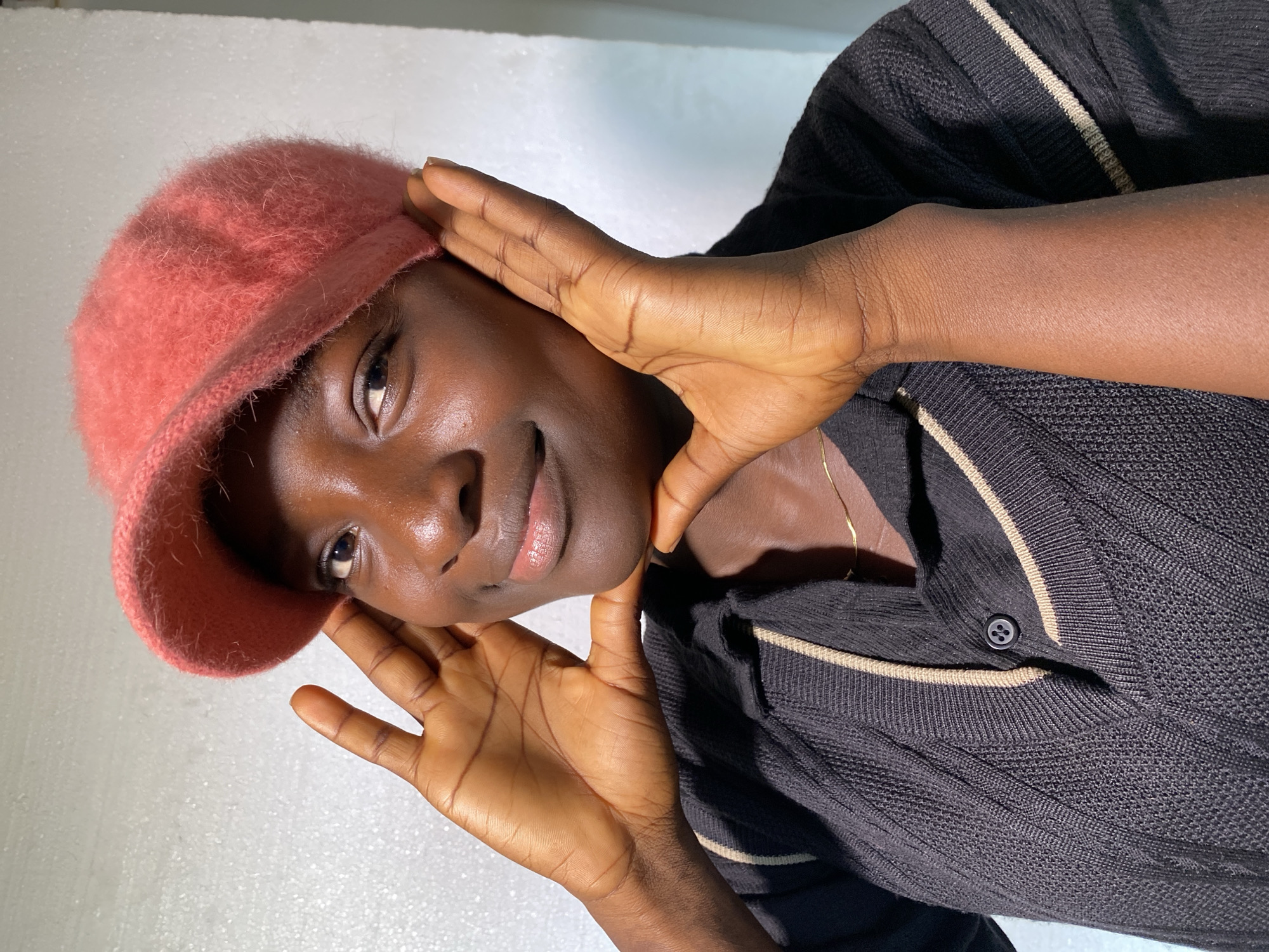 The media file shows a close-up, portrait-style image of a young Black individual wearing a pink knitted cap and a black, ribbed sweater with beige trim. The individual's hands are positioned near their face, with one hand gently touching the side of their head and the other near their chin, creating a frame. The subject has dark skin, a warm complexion, and is looking towards the camera with a slight, gentle smile. Their eyes are dark and expressive, and their lips are slightly parted.
The lighting appears to be artificial, casting subtle highlights on the subject's skin and the texture of the cap and sweater. The background is a plain, textured white surface, suggesting an indoor setting, possibly a studio or a well-lit room. The image is oriented with the subject's head tilted to the left. The overall mood conveyed is one of gentle confidence and quiet engagement. No text is visible in the image. The scene is an intimate portrayal of the individual, focusing on their features and expression.