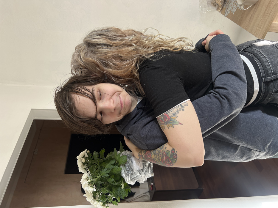 An indoor scene depicts two individuals embracing in what appears to be a residential setting. A male individual, positioned on the left, has medium-length dark brown hair and wears a dark grey hooded sweatshirt. He holds a potted plant of white chrysanthemums, wrapped in clear plastic, in his left arm. His facial expression exhibits a slight frown, and his gaze is directed downwards and to the right. A female individual, largely seen from behind, has long, wavy light brown hair and wears a black short-sleeved top with white trim on the sleeves and dark jeans. She has traditional-style tattoos featuring roses and foliage on her forearms, which are wrapped around the male.
The background on the left shows a light-framed doorway leading into another room with darker walls and flooring, where a dark, indistinct object is partially visible. On the right, a plain light-colored wall is present, along with a partial view of a light wooden table. On this table, a white cup rests on a white snowflake-shaped coaster, next to a clear bag containing light-colored food items. The specific location within Saint Petersburg, Russia, is not discernible from the visual evidence.