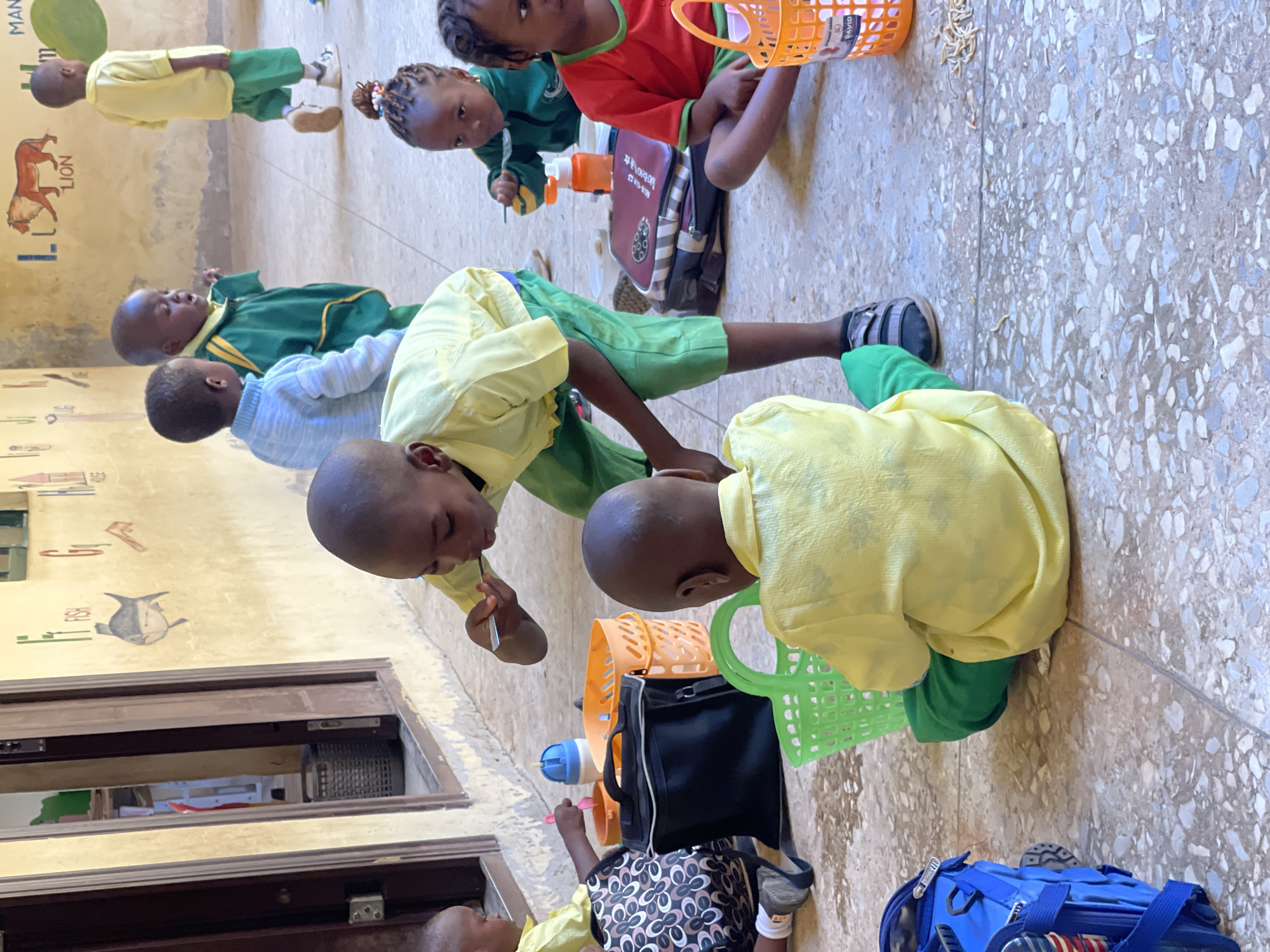 In Akwanga, Nigeria, children are engaged in an outdoor activity, possibly at a school or community center. The ground is paved with speckled concrete tiles, and a cream-colored wall with painted educational murals serves as the backdrop. The murals depict letters of the alphabet with accompanying illustrations, such as a fish for 'F' and a lion for 'L'.
Several children are the focus of the scene. In the foreground, two children are seated on the ground. One child, wearing a yellow shirt and green pants, is facing away from the camera, holding a green plastic basket. Another child in a yellow shirt is bent over, actively eating from a small bowl with a spoon. To their right, a girl in a green jacket and a girl in a red shirt are also seated, with the girl in the green jacket also eating with a spoon. In the background, a boy in a yellow shirt and green shorts walks away from the camera. Another boy in a green uniform stands near him.
Various objects are scattered around the children, including a black bag, an orange basket filled with items, a blue backpack, and a blue and white water bottle. The children appear to be eating, suggesting a mealtime or snack break. The environment seems to be an open, outdoor space adjacent to a building. The lighting suggests it is daytime.