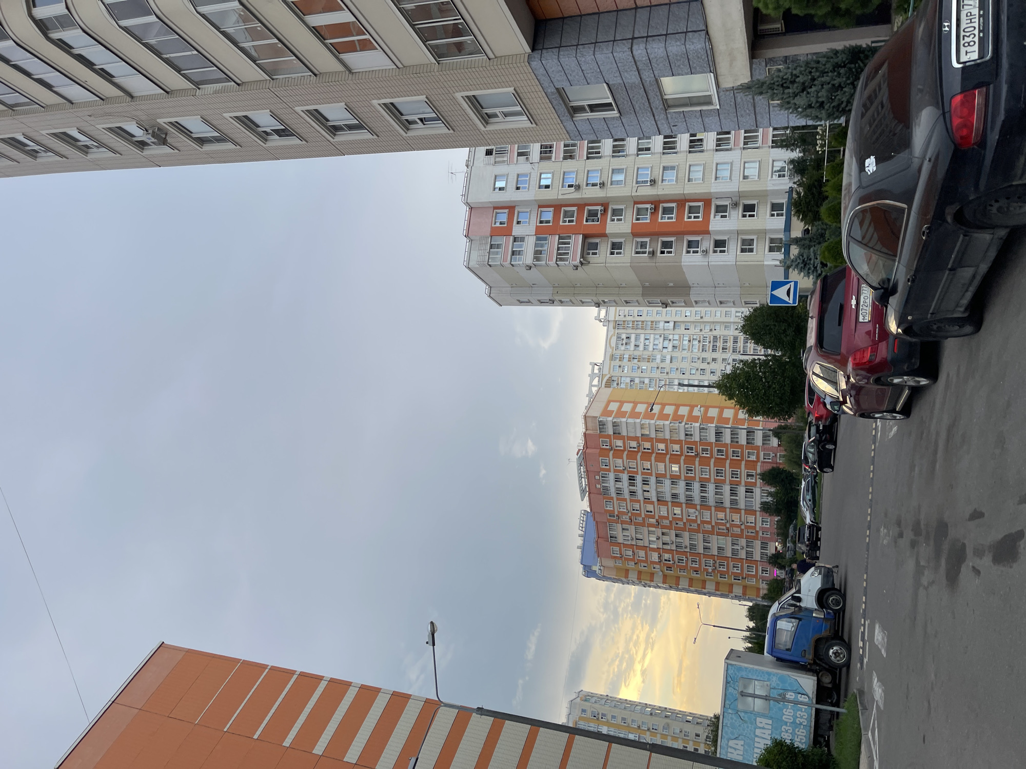 The image depicts a street scene in a residential area of Moscow, Russia, featuring multiple multi-story apartment buildings and parked vehicles. The buildings are characterized by their modern architecture, with colorful facades, numerous windows, and balconies. One building on the left is painted in striking orange and white stripes, while others feature a mix of beige, grey, and orange panels. The sky is overcast with hints of sunset colors visible in the distance.
In the foreground, a blue and white truck with a large boxy trailer is parked on the left side of the street. The trailer has text and graphics on its side. Several passenger cars, including a red SUV and a black sedan, are parked further down the street. A pedestrian is visible walking near the truck. The street surface appears to be asphalt, with some water stains or markings. Green trees and bushes are planted along the sidewalks and around the buildings, adding greenery to the urban environment. A street lamp is visible on a pole near the orange and white building. The overall impression is that of a typical urban residential setting.