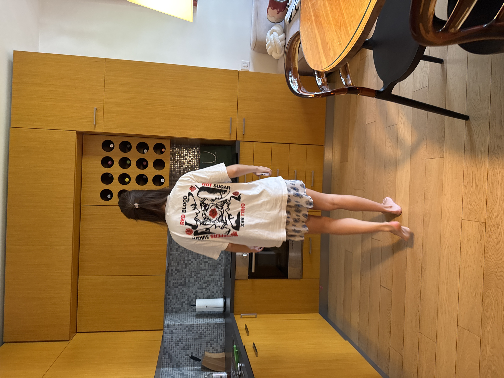 A photograph shows a person standing in a kitchen. The kitchen features light brown wooden cabinetry, including a wine rack. A gray tiled backsplash is visible behind the countertop. A wooden table with black chairs is partially in the frame. The person wears a white t-shirt with a graphic design and patterned shorts. The floor is made of light brown wooden planks.