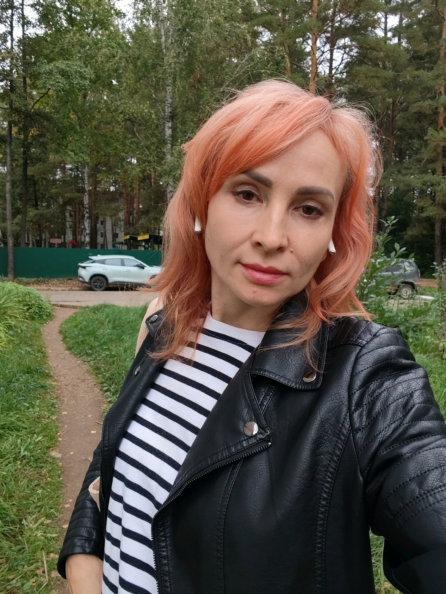 A woman with salmon-pink hair, wearing a black leather jacket over a striped t-shirt, takes a selfie outdoors. She has on white earbuds. In the background, a light-colored car is parked on a paved surface, with a green fence behind it. Further back, buildings and trees are visible. A dirt path with grass on either side is in the foreground. The scene appears to be in a residential area, possibly near a park or wooded area, in Chaikovsky, Russia.