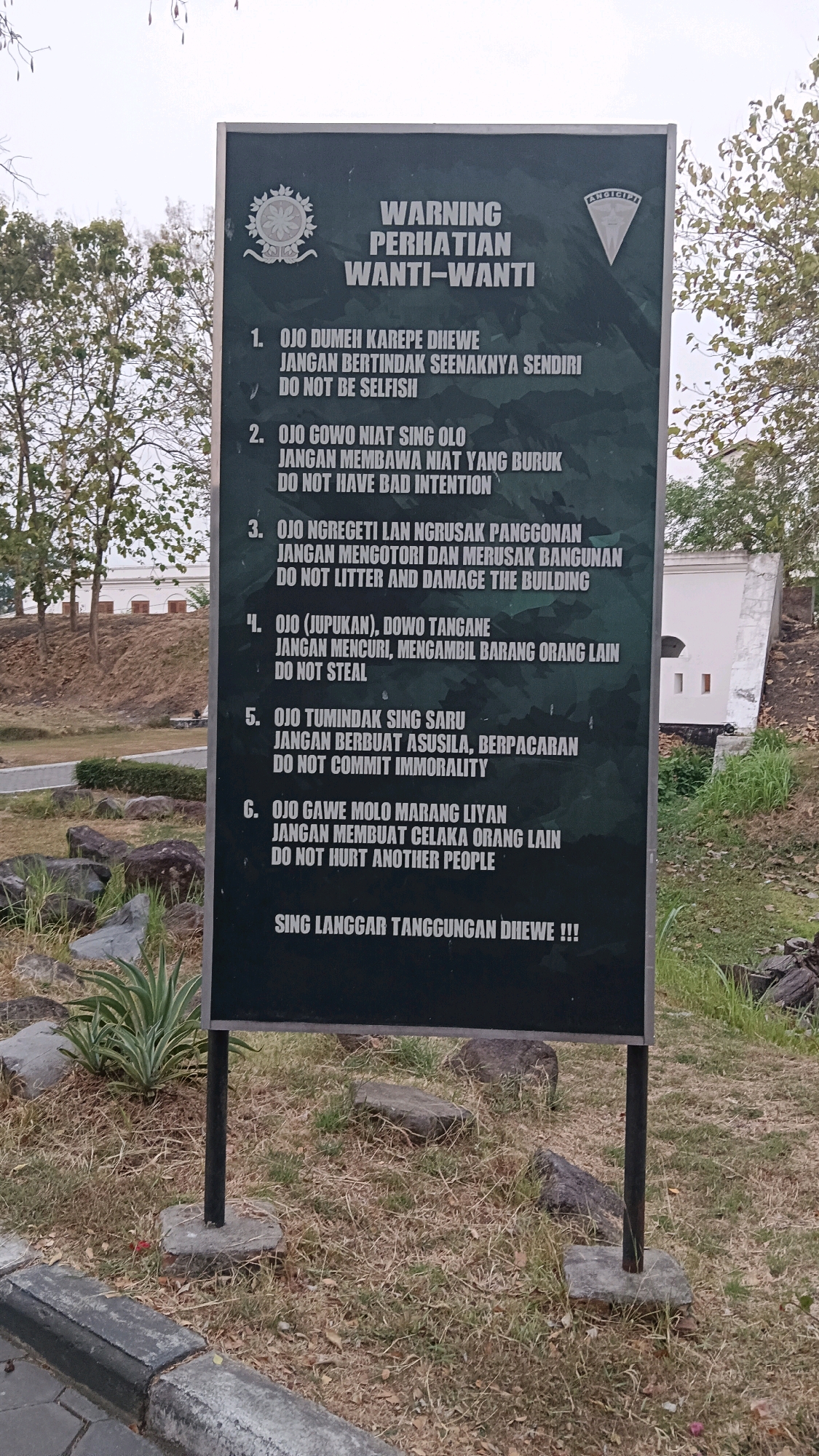 A weathered, dark green sign with white text is mounted on two metal poles, standing upright in a grassy outdoor area. The sign, titled "WARNING PERHATIAN WANTI-WANTI," lists six numbered rules in Javanese and English, along with a concluding sentence in Javanese: "SING LANGGAR TANGGUNGAN DHEWE !!!" The rules advise against selfishness, bad intentions, littering, damaging buildings, stealing, immorality, and hurting others. A circular emblem with intricate designs is visible on the upper left of the sign, and a triangular emblem with the word "ANGICIP" is on the upper right. The sign is positioned on a slight incline with dry grass, scattered rocks, and some greenery at its base. In the background, trees line the left side of the frame, and a partially visible white building with arched openings stands to the right, suggesting a historical or public space. The overall lighting indicates it is daytime, possibly late afternoon, with soft shadows. The sign appears to be located at a historical site in Ngawi, Indonesia, likely related to the building in the background.