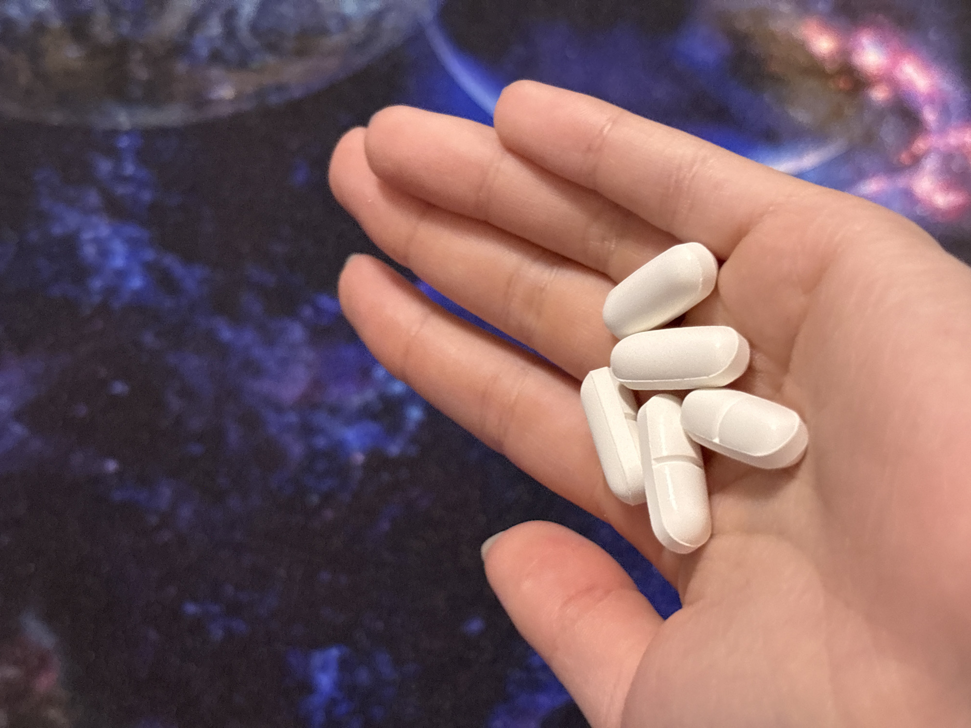 A close-up photograph shows a person's open hand holding several white, capsule-shaped pills with a scored line. The background is a blurred image with patterns of dark blue, brown, and lighter blue and pink, resembling a nebula. The location cannot be determined from the image.
