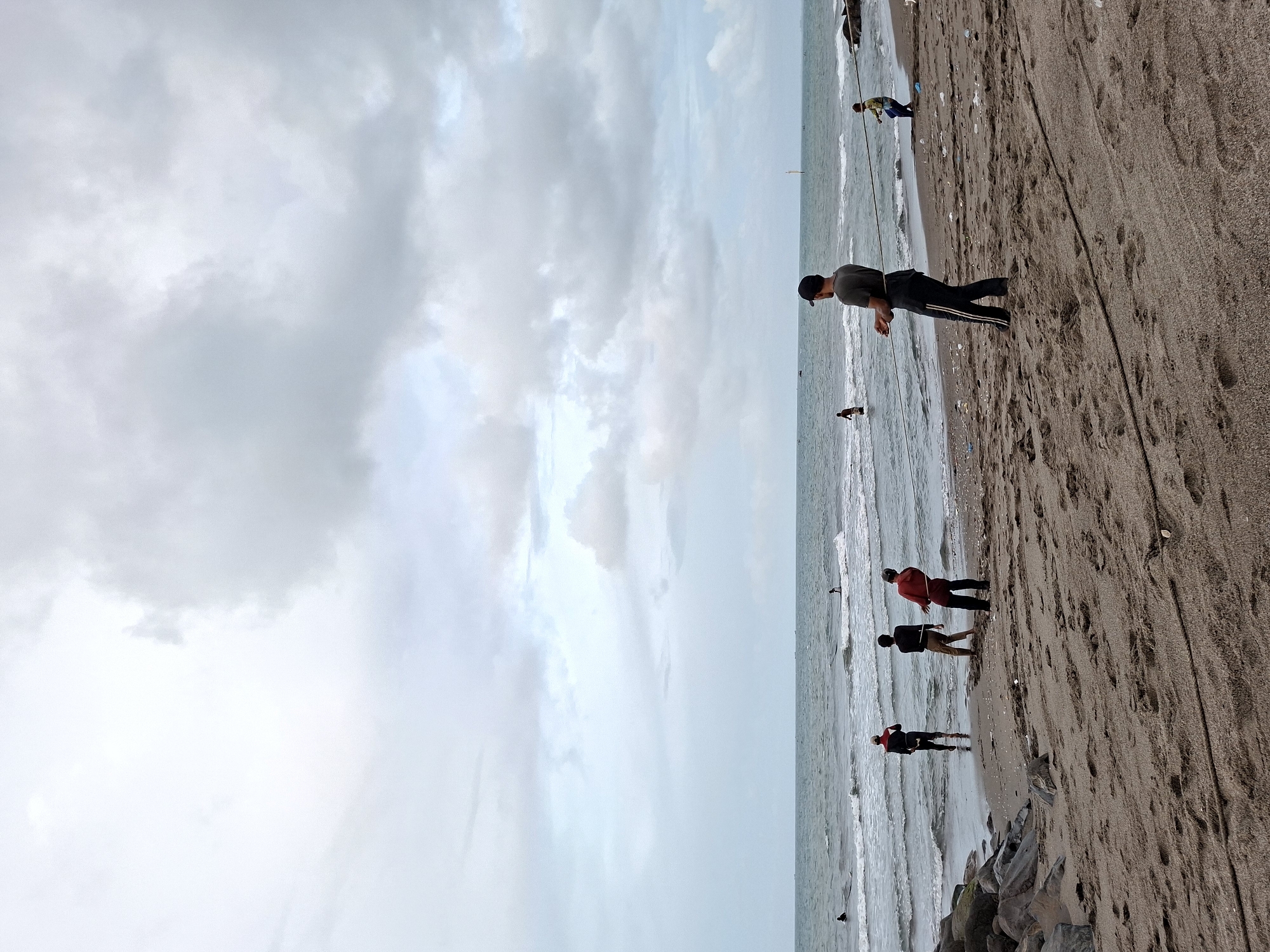 The image shows a beach scene in Lhokseumawe, Indonesia. The sky is overcast with gray clouds. Several people are on the beach and in the water. Near the top of the image, a person is standing on the shoreline holding a long object that appears to be a fishing line. Down the shoreline and farther out into the water, three more people are standing near the water's edge. Towards the bottom of the image, a rocky outcropping is partially visible. The beach sand is dark.