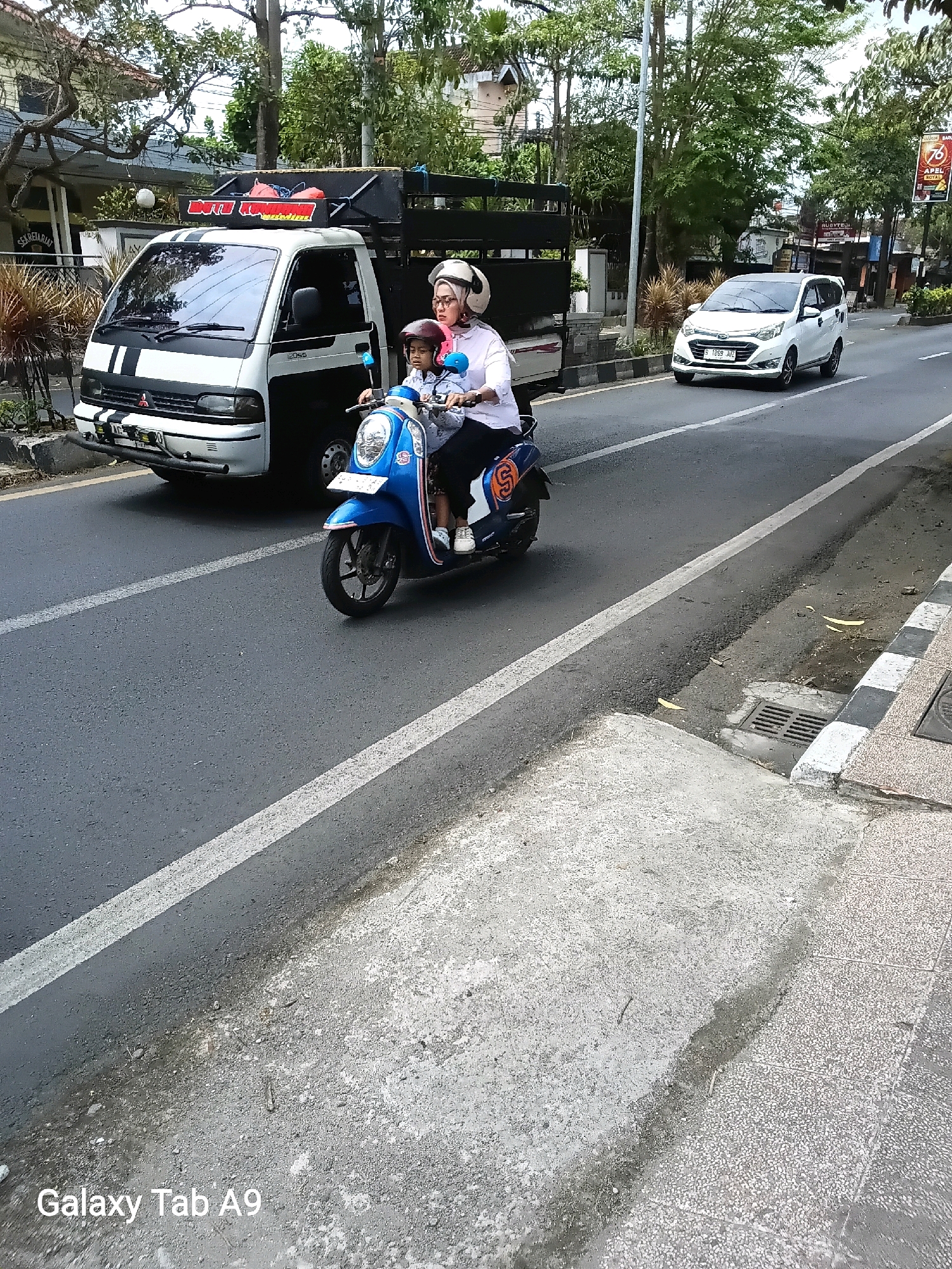 In the city of Kelutan, Indonesia, on a paved road with a white line marking, a woman wearing a white hijab and glasses is riding a blue scooter. She is wearing a white shirt, and a child in a red helmet and blue shirt is sitting in front of her. The scooter has a white stripe with the letter "S" on its side. Behind the scooter, a white truck with black stripes on its hood and a black cargo bed is visible. The truck has a sign with red lettering and an image on its cargo bed. To the right of the truck and scooter, a white car is driving on the same road. The environment includes trees, buildings, and a sidewalk with a drain grate. The text "Galaxy Tab A9" is visible in the lower left corner of the image.