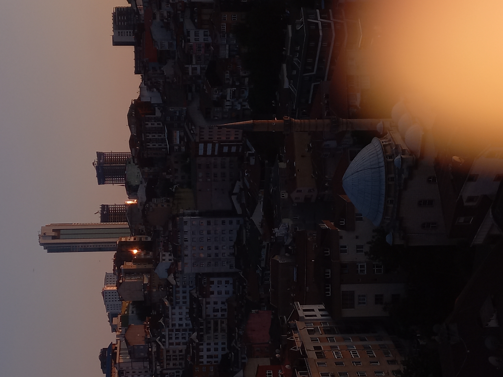 The image depicts a cityscape at dusk, with buildings of varying heights and architectural styles dominating the scene. The sky is a muted gradient of pink and orange, suggesting either sunrise or sunset. In the background, a tall, modern skyscraper stands out with its glass facade reflecting the ambient light. Surrounding it are lower-rise residential buildings, many with visible windows, some illuminated, indicating occupancy.
A notable structure in the mid-ground is a mosque with a domed roof and a slender minaret. The mosque's architectural features, such as the patterned stonework on the minaret and the arched windows, are discernible. The buildings are densely packed, typical of urban environments. In the foreground, a bright, out-of-focus orange glow obscures a portion of the scene, likely lens flare or an intentional photographic effect.
The overall impression is of a lively, inhabited urban area, bathed in the soft, diffused light of twilight. The scene is devoid of any visible people or overt actions. The location is identified as Kâğıthane, Turkey, a district known for its blend of historical and modern architecture, and its residential character.