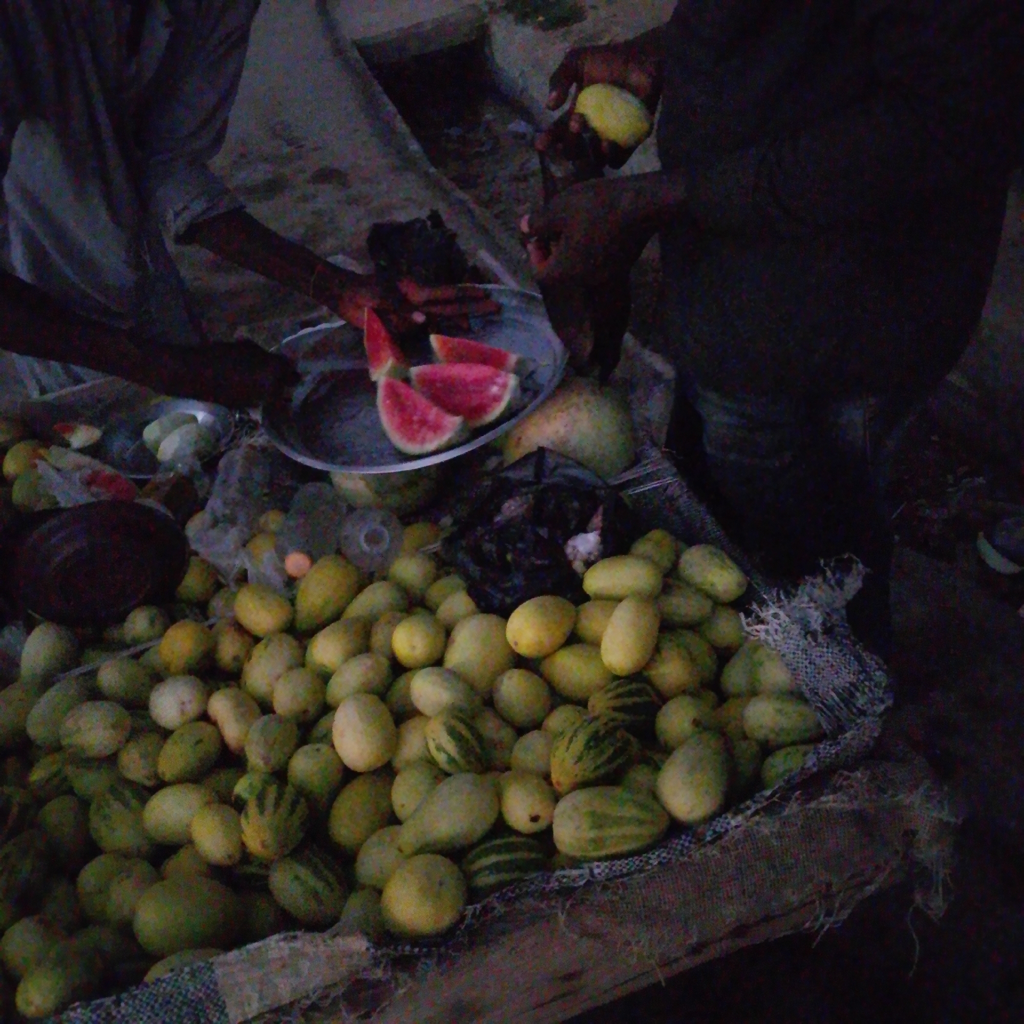 In Wuro Allahamdu, Nigeria, a vendor, partially obscured by shadow, is seen preparing fruit. Sliced watermelon rests on a metal plate, while other fruits, possibly melons or gourds, are piled in a sack. The ground appears to be dirt. The scene suggests an outdoor market or roadside stall, illuminated by dim, possibly artificial, light.