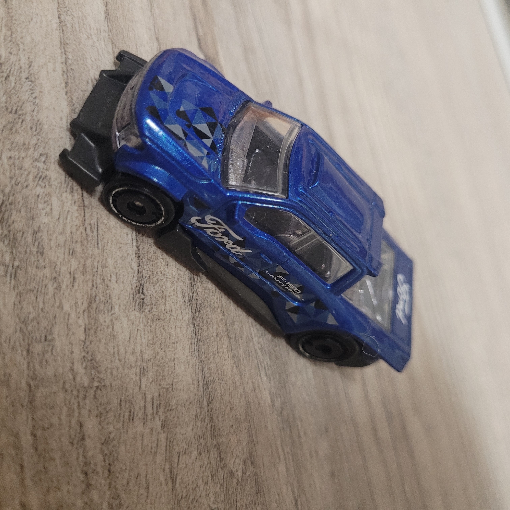 A blue Ford F-150 Lightning toy truck is photographed from a high angle, angled to the left, on a wooden surface. The toy truck is detailed with "Ford" branding on the side and hood, and the "F-150 LIGHTNING" text on the door. The truck has black wheels and a black undercarriage. The background is a light brown wooden surface with a visible grain pattern.