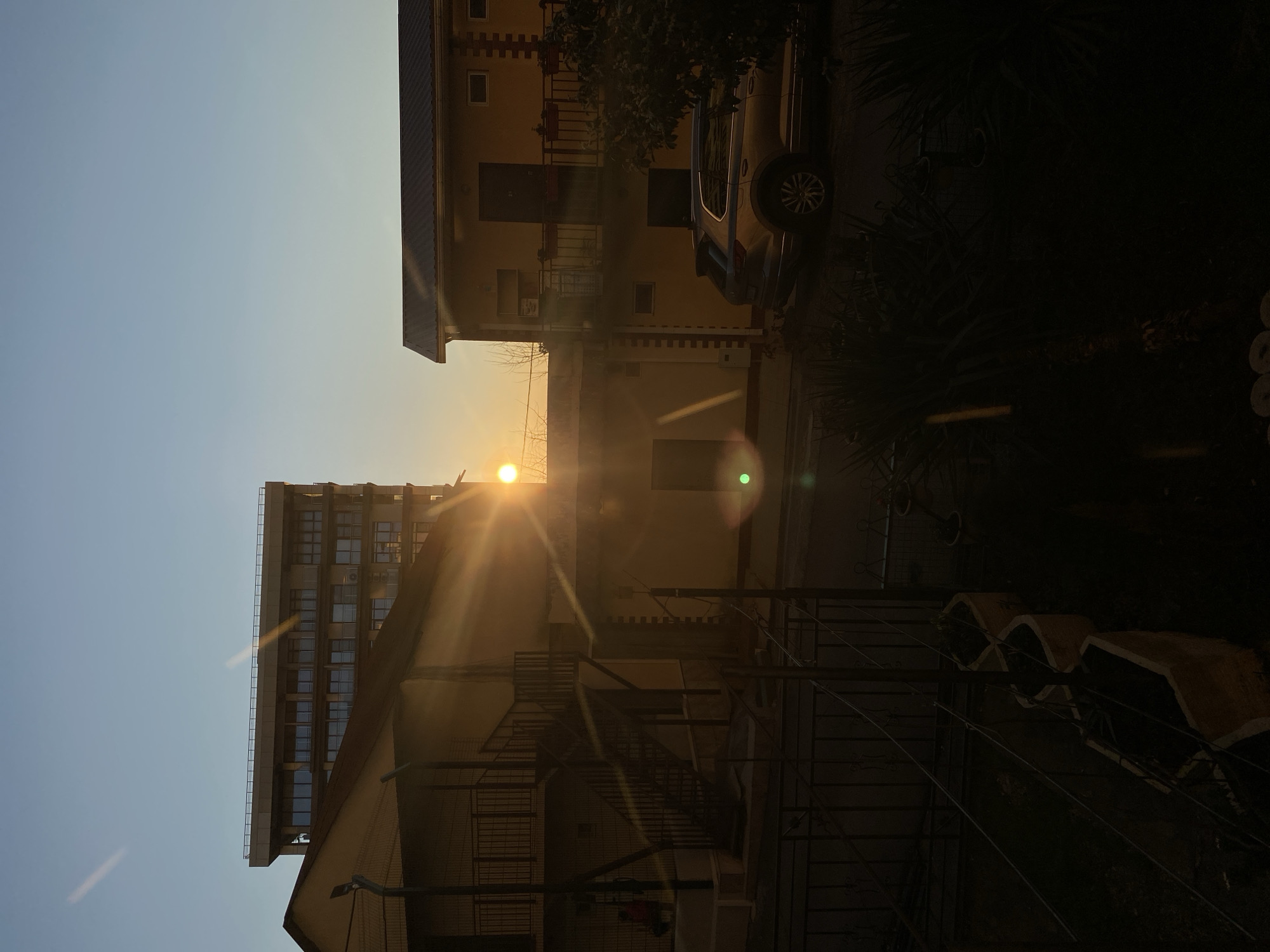 The image shows a scene in what appears to be the city of Sochi, Russia, with buildings and a car visible. The sun is low in the sky, creating a bright glare. The architectural styles suggest a mix of old and new buildings. The photograph shows a partly-obstructed view of residential and commercial structures with a car parked beside one building. There are trees and some other vegetation. The view is urban, with a lot of concrete and masonry buildings.