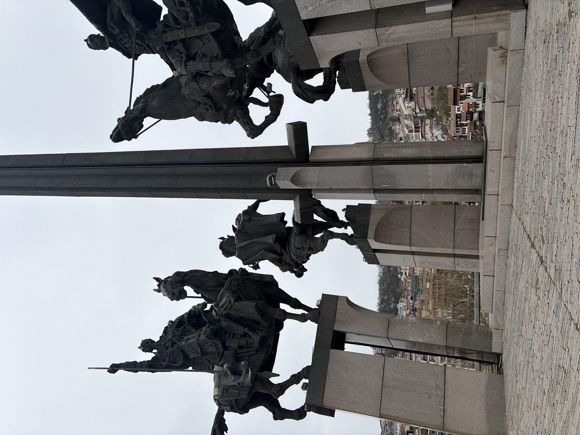 The image shows a monument in Veliko Tarnovo, Bulgaria. The monument consists of three groups of sculptures depicting mounted horsemen. The sculptures are made of dark metal or stone, set against a backdrop of a cloudy sky and cityscape. The monument sits on a stone platform. The city of Veliko Tarnovo is visible in the background.
