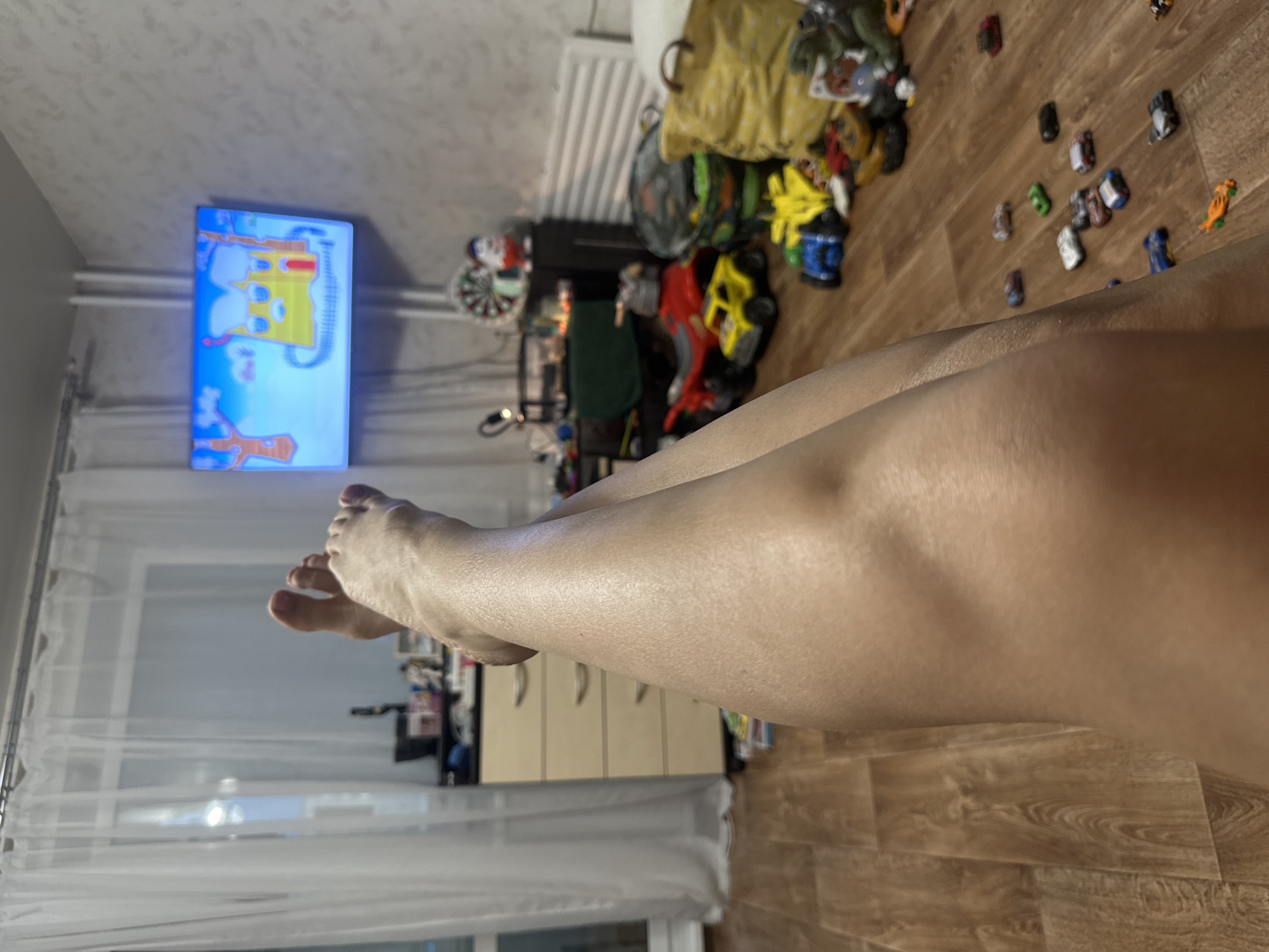 The image depicts a domestic indoor scene, likely a living room or playroom, located in Novoaltaysk, Russia. The foreground is dominated by a close-up view of a person's legs and feet, with the camera positioned low and looking upwards. The legs are crossed and appear to be resting. In the background, a television screen displays a cartoon with a yellow house and trees. The room features a dresser with drawers, a window with sheer white curtains, and a wall with textured wallpaper. Scattered on the wooden floor are various children's toys, including toy cars of different colors and sizes, a toy motorcycle, a dartboard, and a large yellow fabric bag. A white radiator is visible against the wall. The overall impression is of a casual, home environment where a person is relaxing while watching television, with a collection of children's toys present in the vicinity.