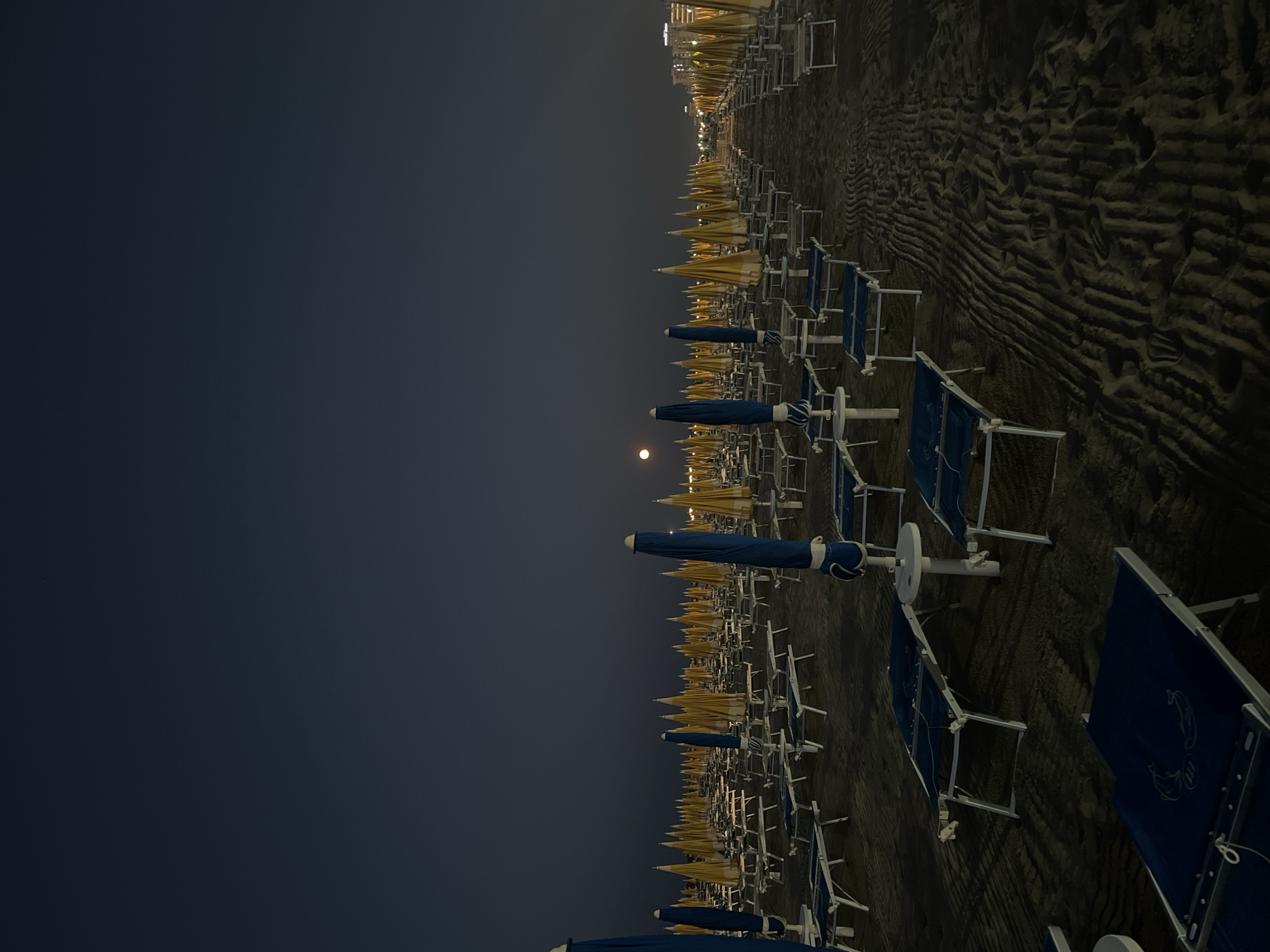 An empty beach in Rimini, Italy, is depicted at dusk or night, with rows of closed beach umbrellas and lounge chairs stretching into the distance. A pale moon is visible in the dark sky. The sand shows ripples from waves or wind, and some footprints are present. The scene is quiet and still, suggesting the end of a day at the beach. Buildings are faintly visible in the far distance, illuminated by lights.