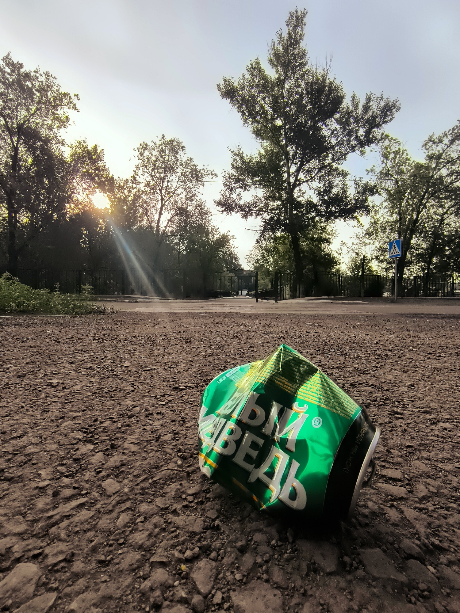 A crumpled, green aluminum can lies on a gravelly surface in front of a paved area. The can is positioned on the lower right side of the frame and shows Cyrillic lettering, primarily white with some yellow accents. Behind the can, trees line the edge of the paved area, with the sun breaking through the foliage in the upper left quadrant, casting sunbeams onto the scene. Further in the background, a pedestrian crossing sign is visible on the right, and a metal fence runs along the treeline. The sky is bright and overcast. The location is the city administration area in Qarağandı, Kazakhstan.