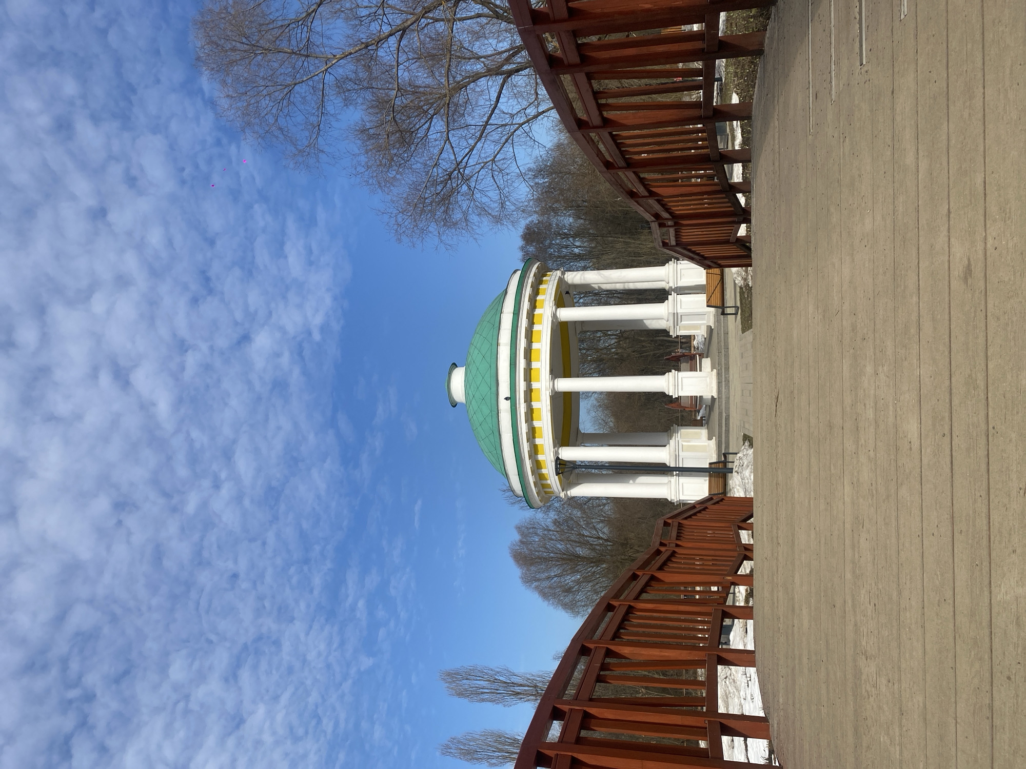 An outdoor scene captured from a low vantage point depicts the Gagarin's Rotunda in Vorontsovo Estate Park, Moscow, Russia. In the immediate foreground, a weathered wooden plank bridge extends towards the rotunda, flanked by reddish-brown wooden railings that curve inward. The rotunda features a prominent green dome with a small finial, a yellow frieze, and a base of white classical columns, standing on a grey paved area. Two dark benches are visible near the rotunda's base, one on each side, along with a black light pole on the left. Patches of residual snow are present on the ground beside the bridge and near the rotunda. The background consists of numerous bare deciduous trees and dense woods. The sky above is clear blue with scattered white, patchy clouds. No individuals are present in the frame.