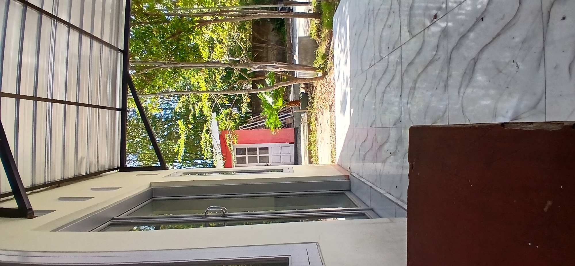 The image shows a sunlit outdoor patio area with a tiled floor. To the left, a cream-colored building features a large glass sliding door with a silver handle. Above the building's wall, a metal-framed corrugated metal roof extends outwards, casting shadows on the patio. Beyond the patio, a small, pink outbuilding with a white-paneled door and a white trim is visible. Several trees with green foliage are present in the background, along with some vegetation and what appears to be a concrete structure. The scene is set during the daytime.
