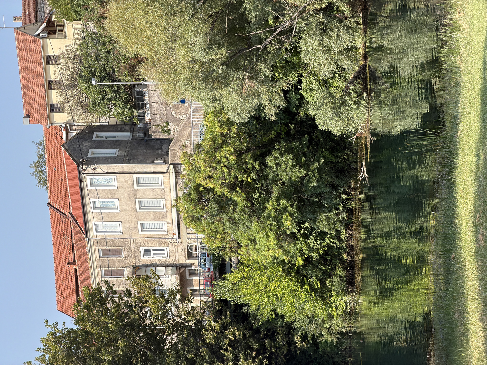 The image depicts a serene waterscape in Trebinje, Bosnia and Herzegovina, with a focus on the Trebišnjica River and the surrounding lush greenery. In the background, traditional residential buildings with red tiled roofs are visible, perched on a hillside. One building, painted a pale yellow, features wooden shutters and a balcony. Adjacent to it is a grey building with multiple windows. The buildings are partially obscured by dense foliage from mature trees, their branches reaching towards the structures.
A bridge or elevated walkway runs across the middle ground, with two cars, one red and one white van, parked on it, suggesting vehicular access. The foreground is dominated by the tranquil surface of the Trebišnjica River, reflecting the vibrant green of the trees and the blue of the sky. The water is calm, with gentle ripples distorting the reflections of the overhanging vegetation. The riverbank on the near side is lined with lush green grass. The overall atmosphere is one of natural beauty and quietude, characteristic of the idyllic setting in Trebinje.