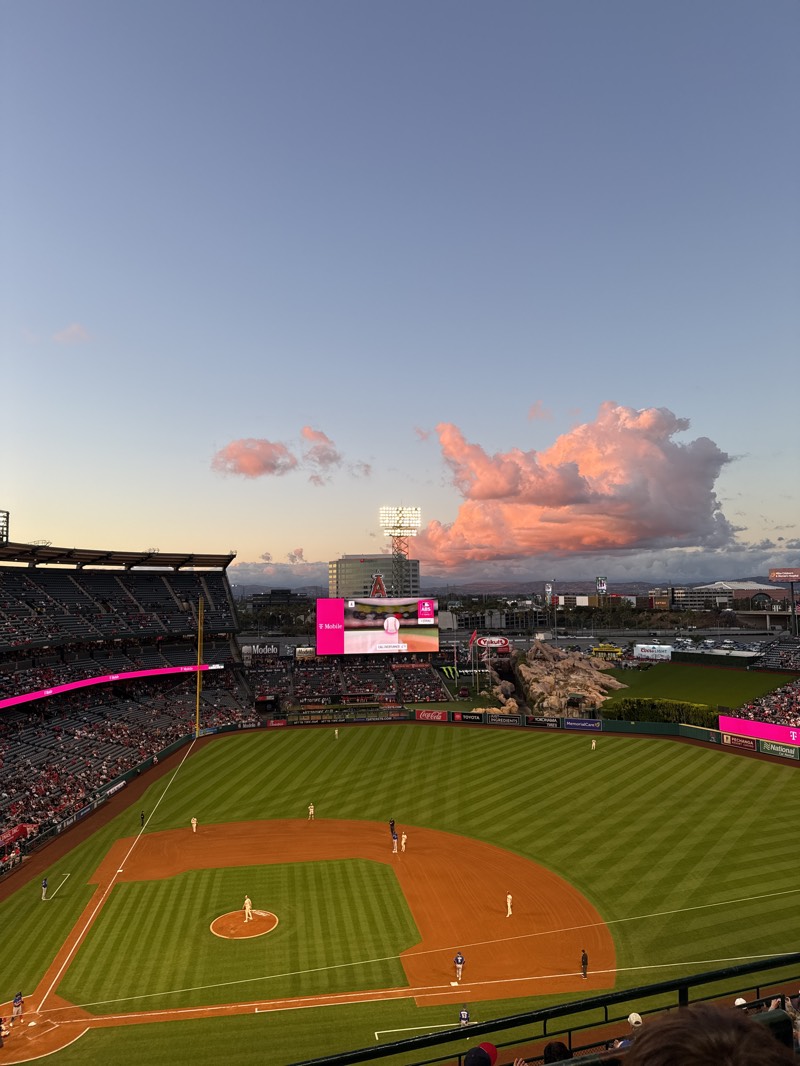 The image captures an elevated view of a baseball game in progress at Angel Stadium in Anaheim, United States, during sunset. The sky displays a serene gradient from light blue overhead to warm pink and orange hues on scattered cumulus clouds, illuminated by the setting sun. Stadium lights are already on, casting a glow over the scene.
The central subject is the baseball field, with its meticulously striped green grass outfield and infield, contrasting with the reddish-brown dirt of the baselines and pitcher's mound. Several baseball players are visible on the field, including a pitcher on the mound, a batter at home plate, and fielders positioned around the bases, indicating an active game. The multi-tiered stadium seating is filled with numerous spectators, particularly on the first base side (left of the image), suggesting a well-attended event.
Notable stadium features include a large video scoreboard in the center-left outfield displaying a baseball graphic and the "T-Mobile" logo. To its right, a distinctive rock formation resembling mountains, a signature element of Angel Stadium, is visible behind the outfield wall. Various advertisements line the outfield fence and are visible on screens and buildings. Text visible includes "Angels" on a building in the background, "T-Mobile," "Modelo," "Yakult," "Coca-Cola," "Toyota," "MemorialCare," "National," and "Pechanga Resort Casino" on a smaller screen to the right. The overall impression is a vibrant evening at a professional baseball game under a striking sunset.